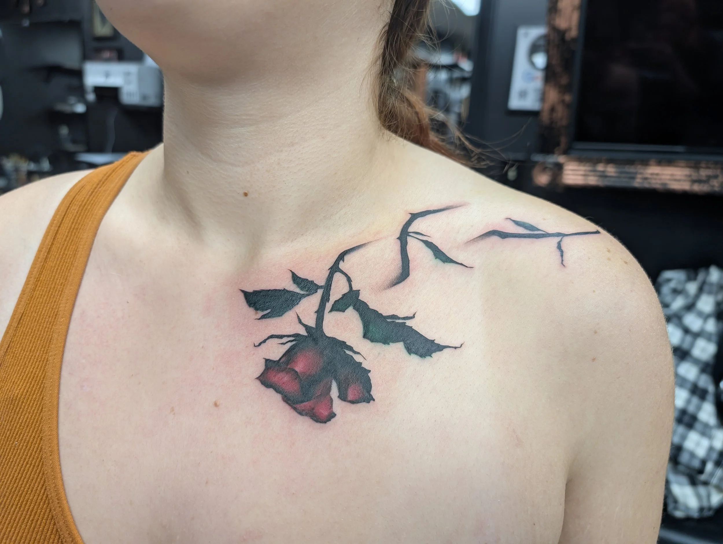Close-up of a person's shoulder and neck showing a black tattoo of a branch or tree with leaves, partially visible, on fair skin. The person is wearing an orange sleeveless top. Background features blurred objects, possibly in a salon or barber shop.