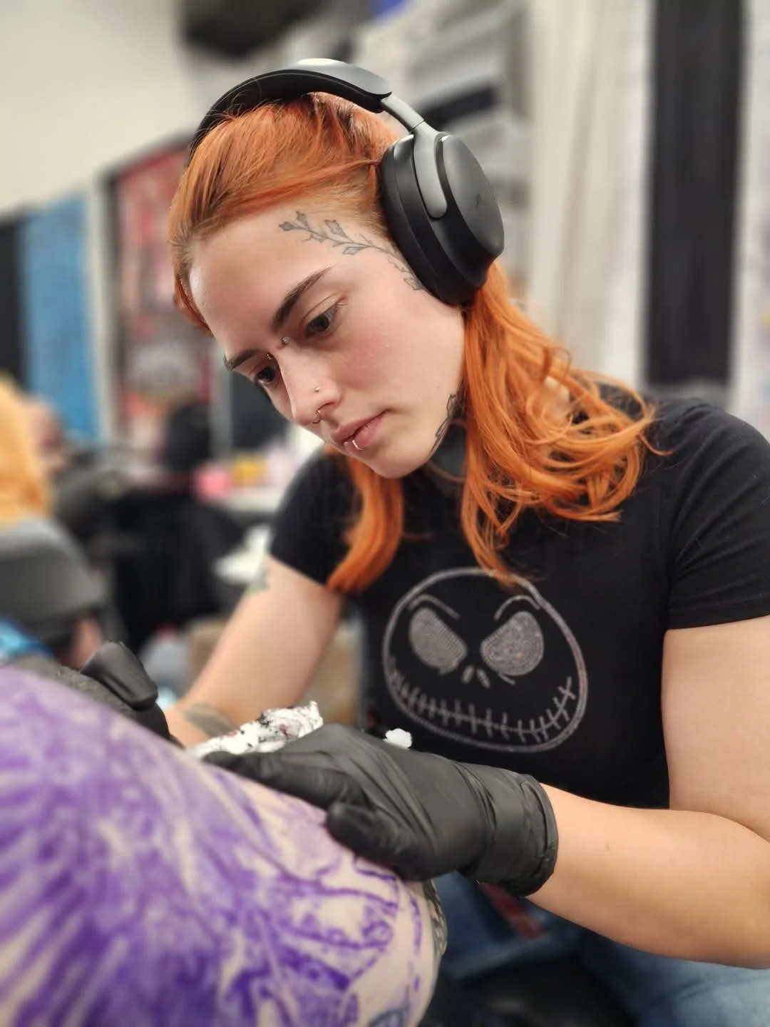 Tattoo artist with orange hair, wearing black gloves, black headphones, and a black t-shirt with a skull design, tattooing a person's arm in a tattoo studio.