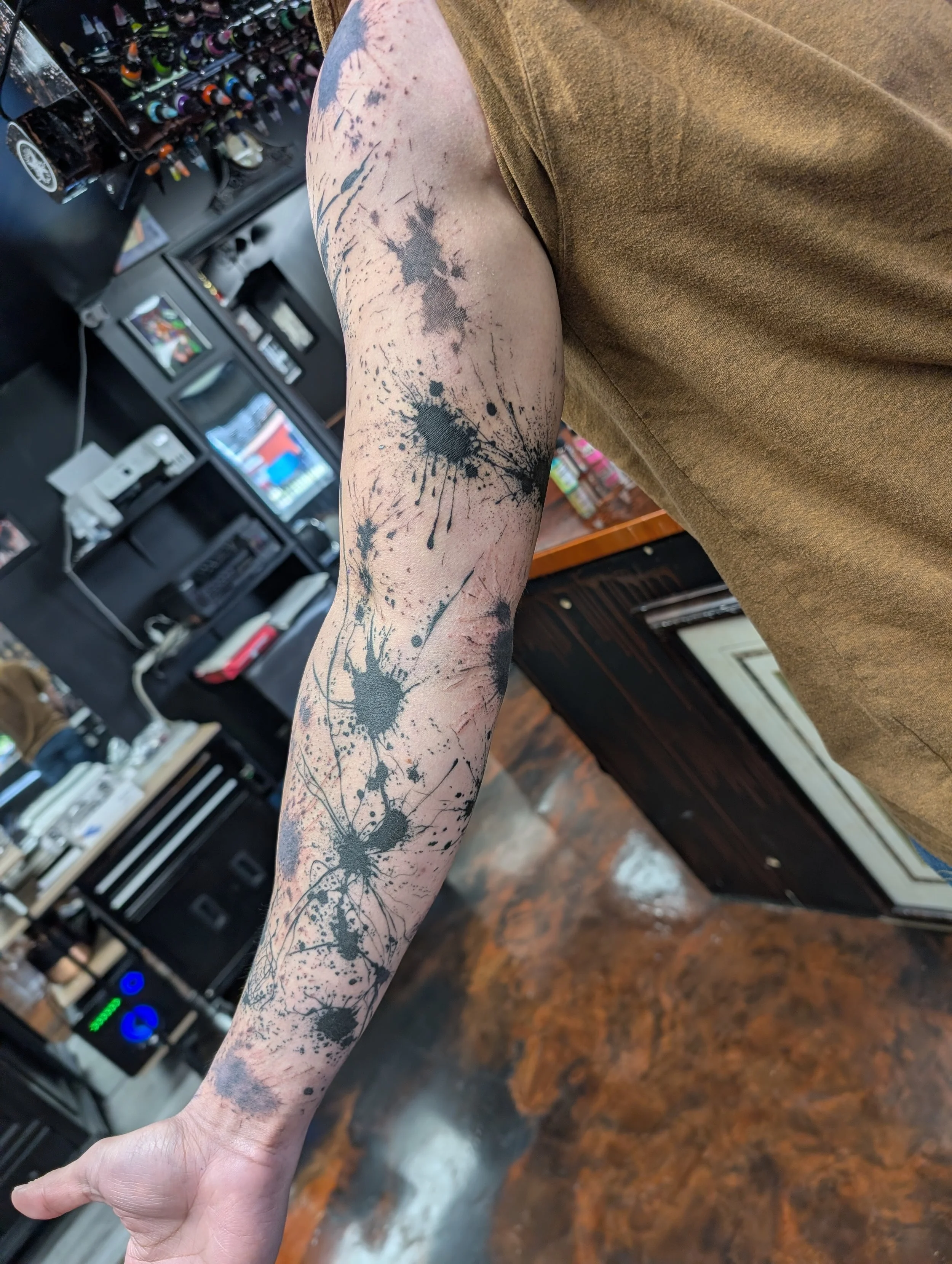 A person's arm with a tattoo of black ink splatters and splashes, in a style resembling paint splatter or ink blot. The arm is resting on a brown wooden surface, and background elements include electronic equipment, a soda or juice machine, and a she