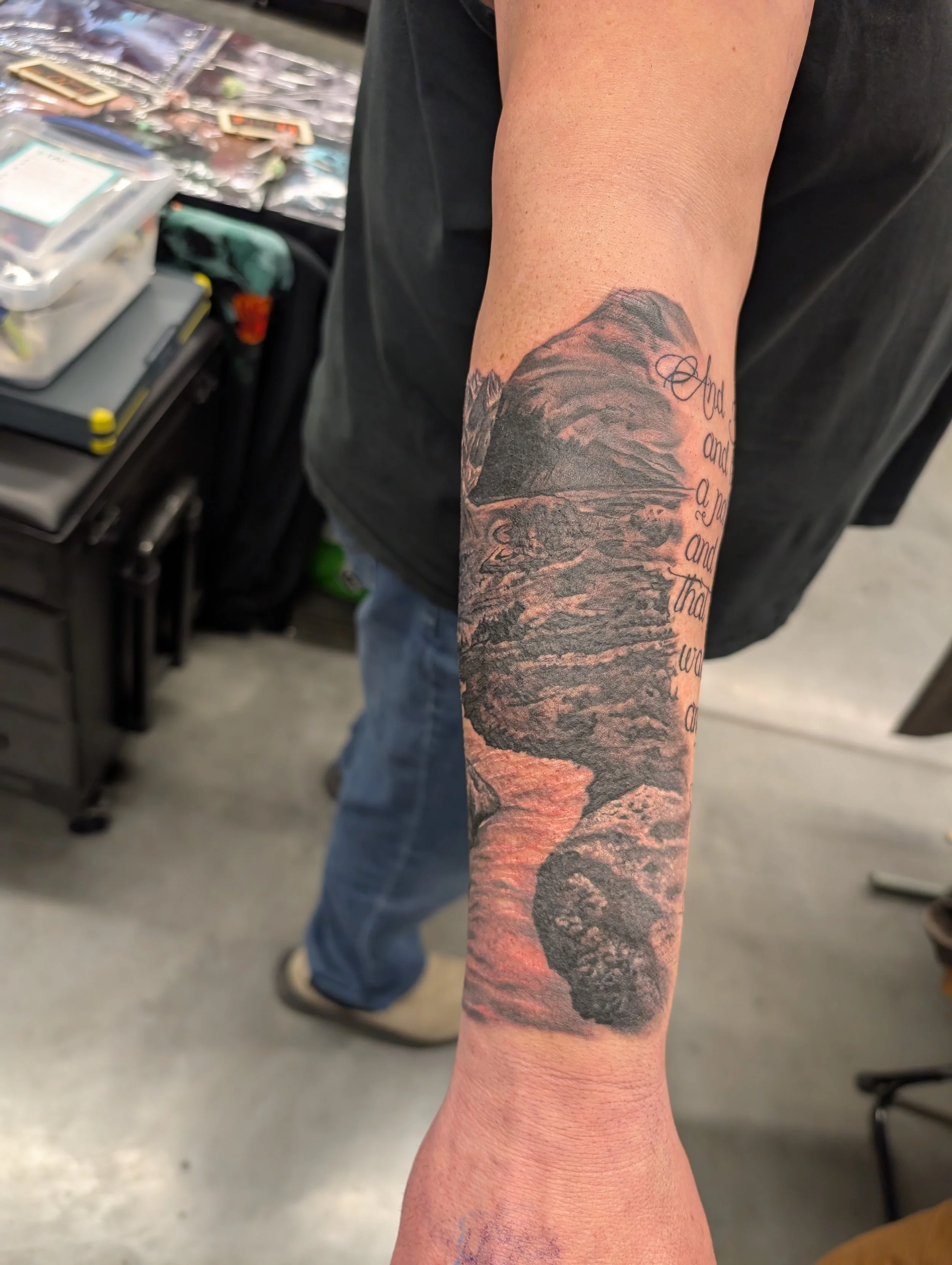 Close-up of a person's arm with a detailed tattoo of a mountain landscape, including mountains, rocks, and water, along with cursive text. The background shows tables with various tattoo supplies and a workspace setting.