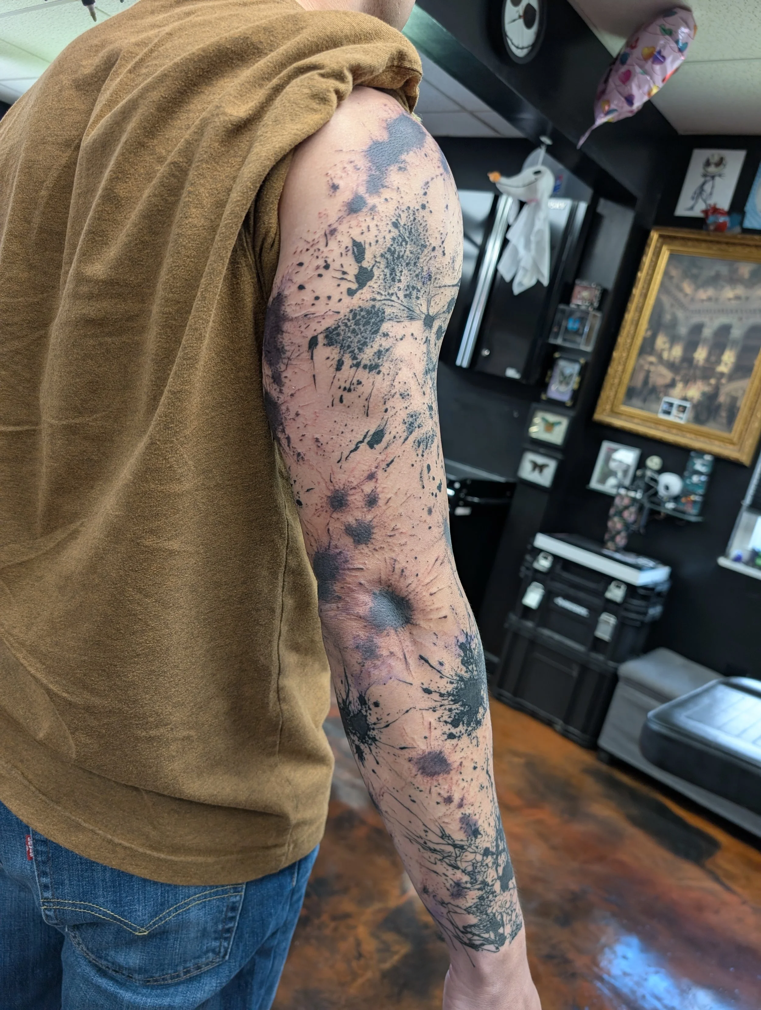 Person with a large black and gray tattoo sleeve on their left arm, wearing a brown short-sleeved shirt and blue jeans, standing indoors with black shelving in the background and framed artwork on the wall
