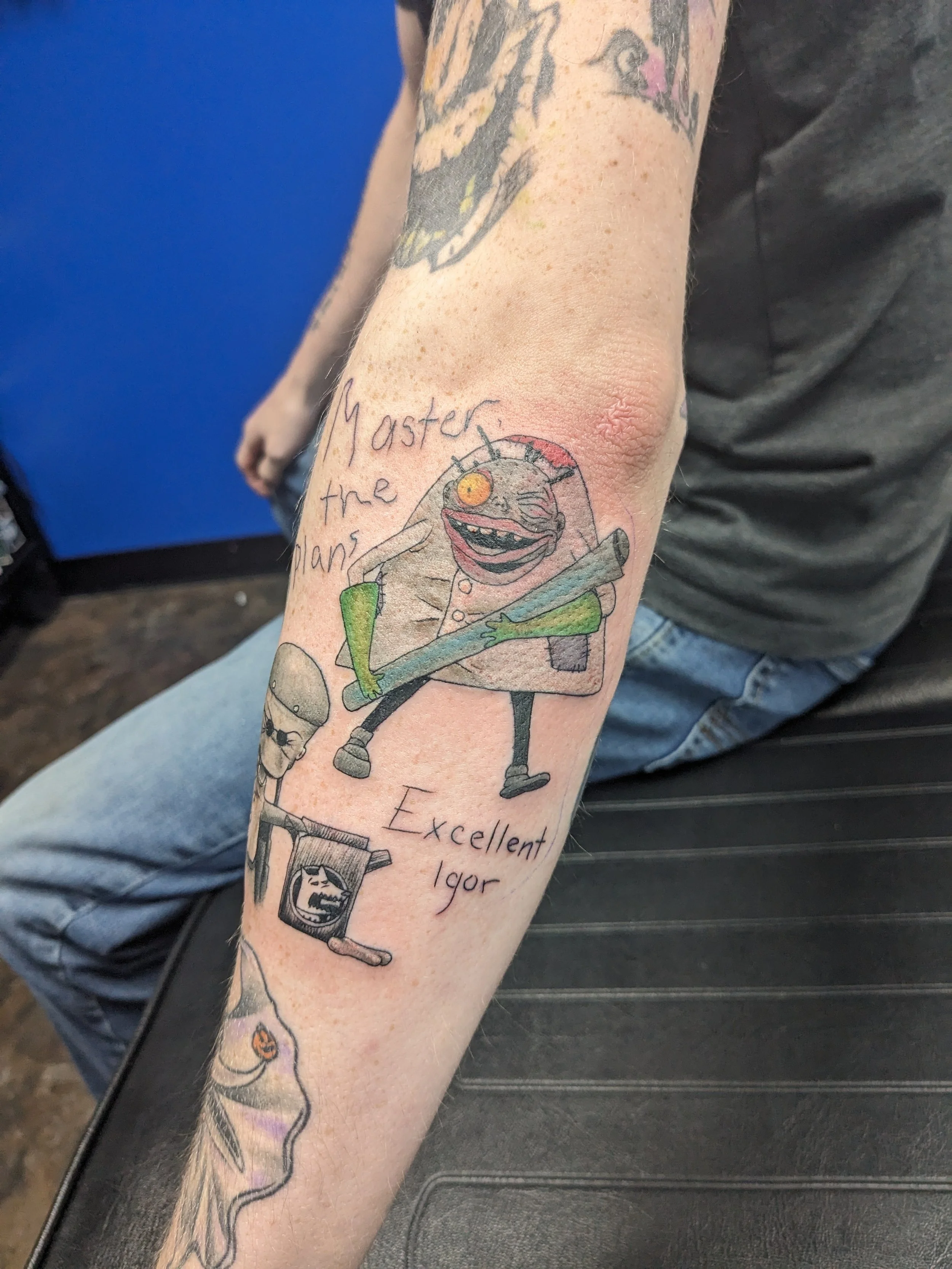 Tattoo on person's arm featuring a grinning cartoon character with a spiky hairstyle, holding a baseball bat, with the handwritten words 'Master the Man's' and 'Excellent Igor' around it.