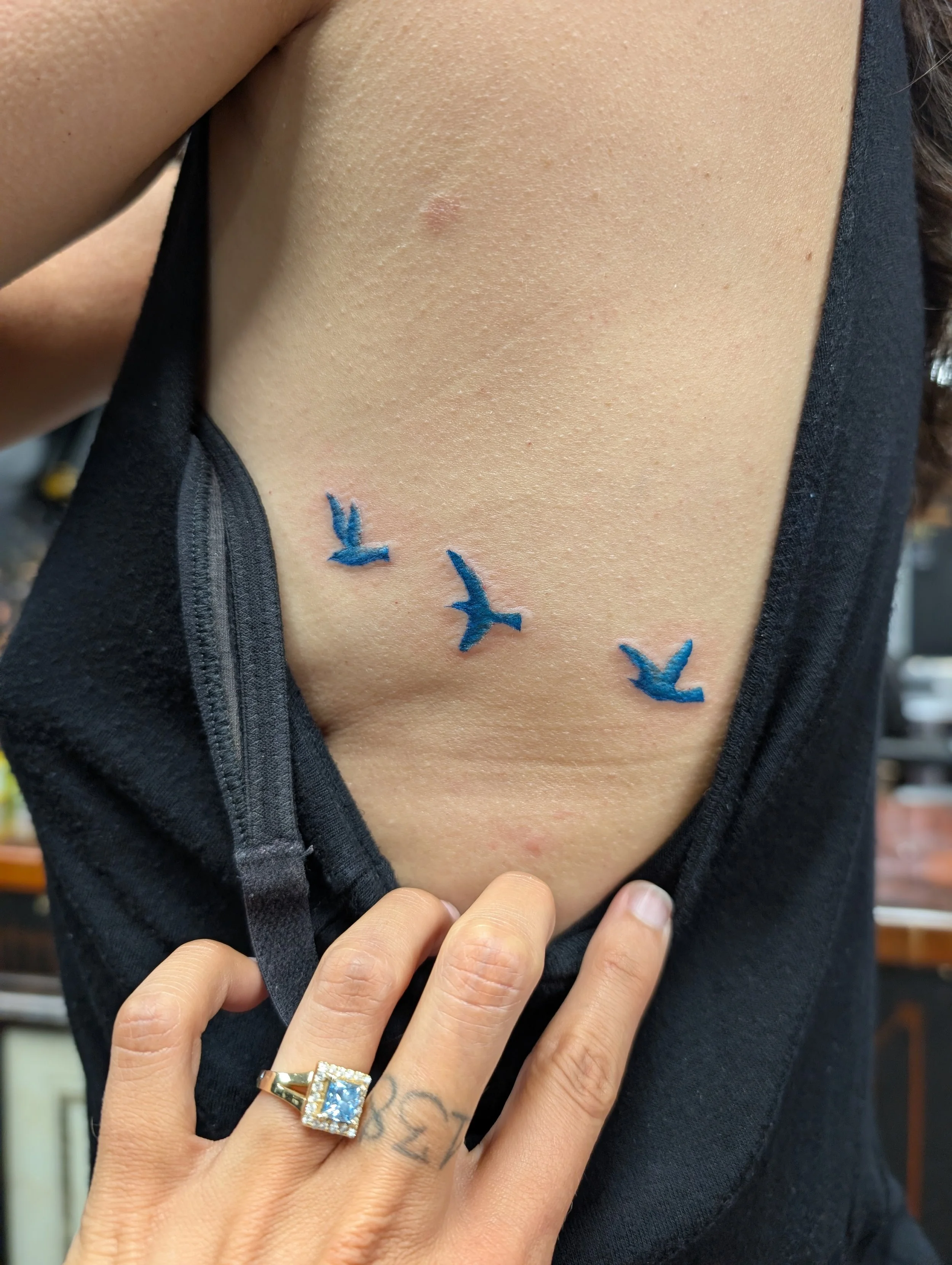Close-up of a person's chest and hand, showing a tattoo on the person's finger and a large ring with a blue gemstone. The person is wearing a black top with a partially unzipped zipper.