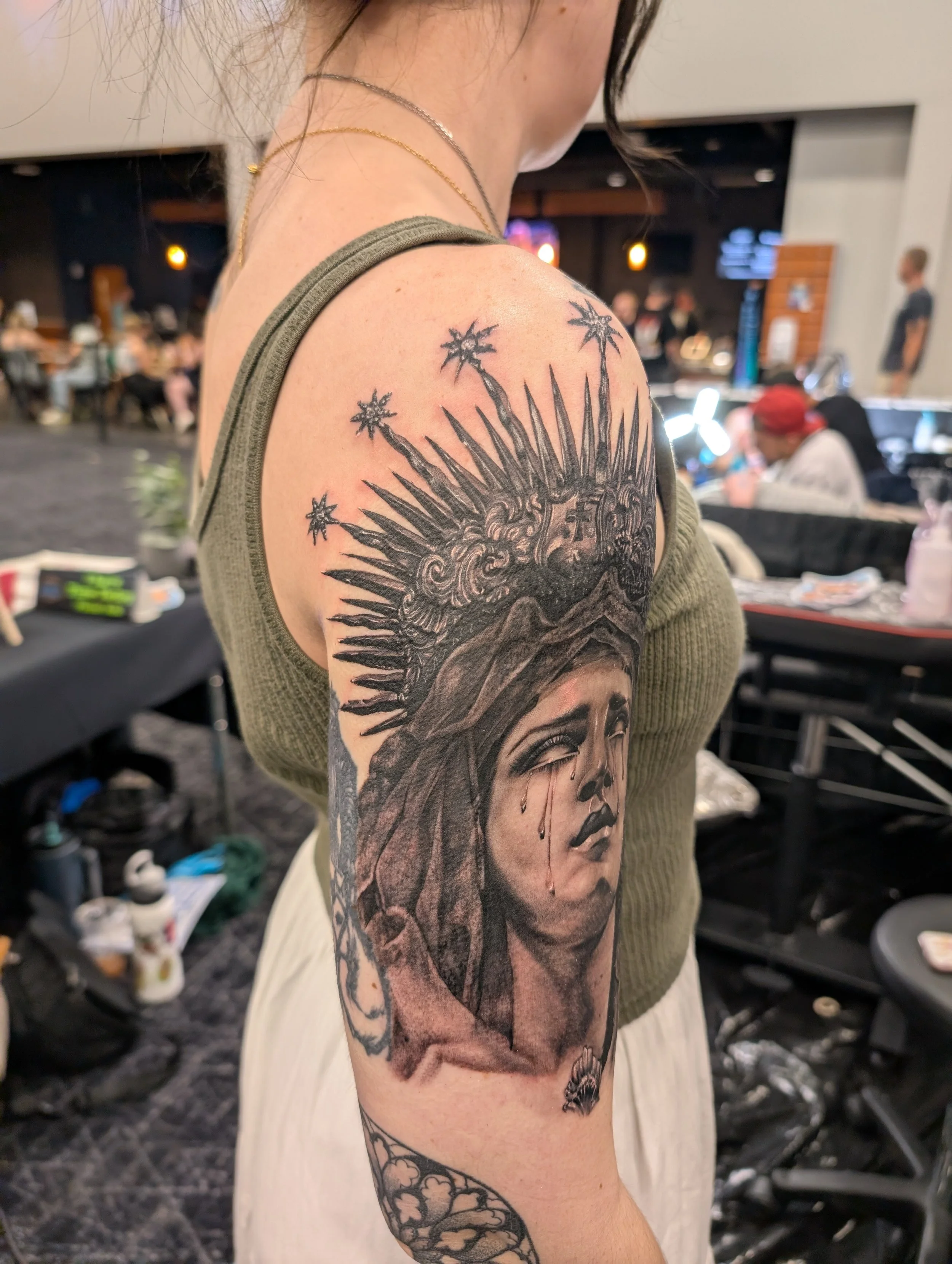 Close-up of a person's shoulder with a detailed black tattoo of a woman's face, with tears running down her cheeks, and a crown of thorns on her head. The background shows a busy indoor setting with people, tables, and chairs.