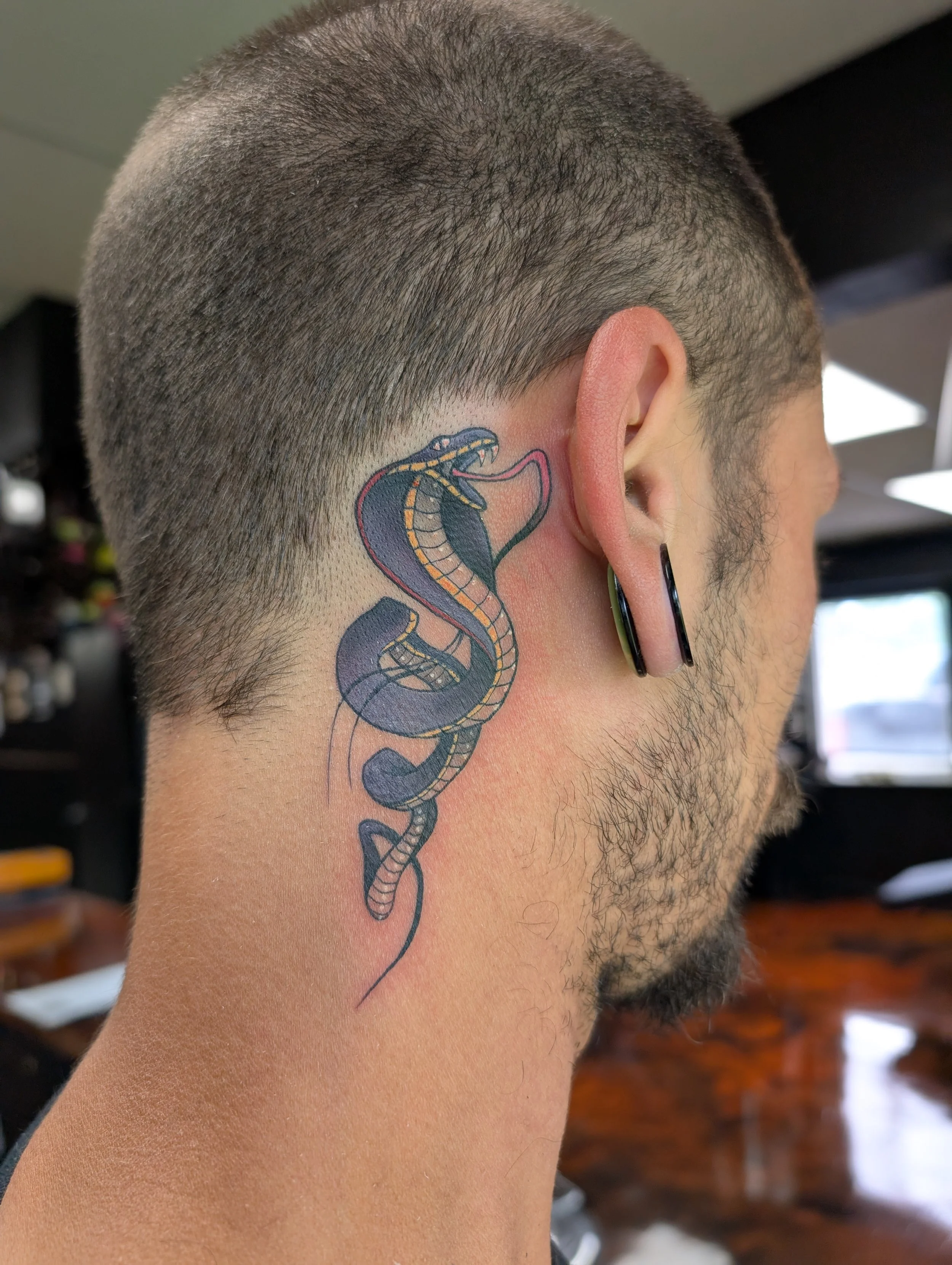 Close-up of a man's head showing a snake tattoo behind the ear, with short hair and ear piercings.