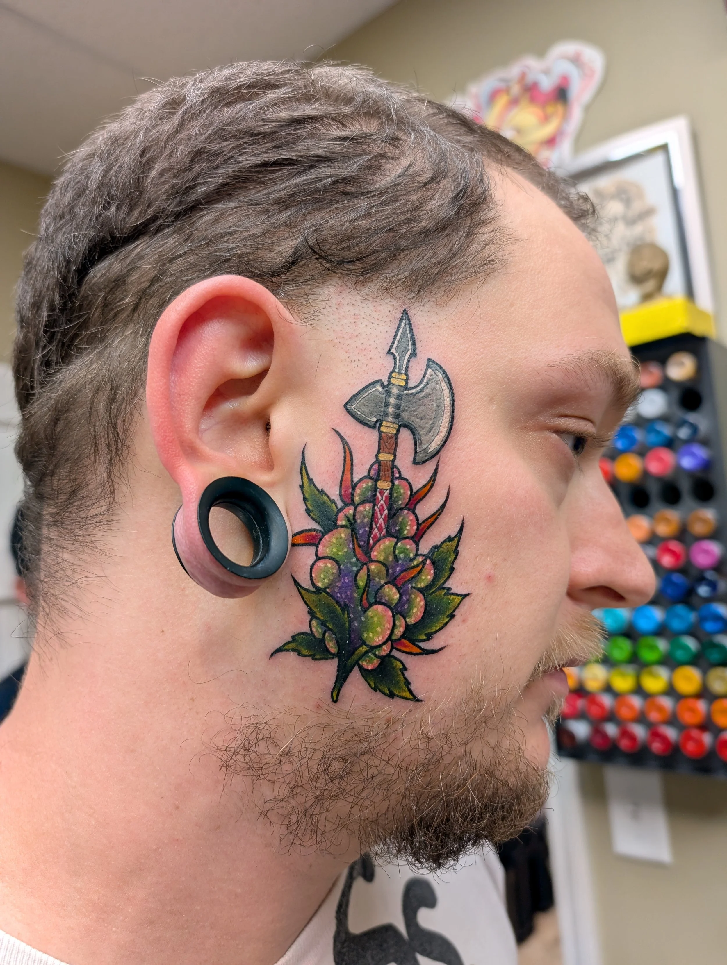 Close-up of a man's face showing a tattoo of an axe with a decorated handle on his cheek, with colorful flowers below it. The man has brown, curly hair and large black ear gauges.