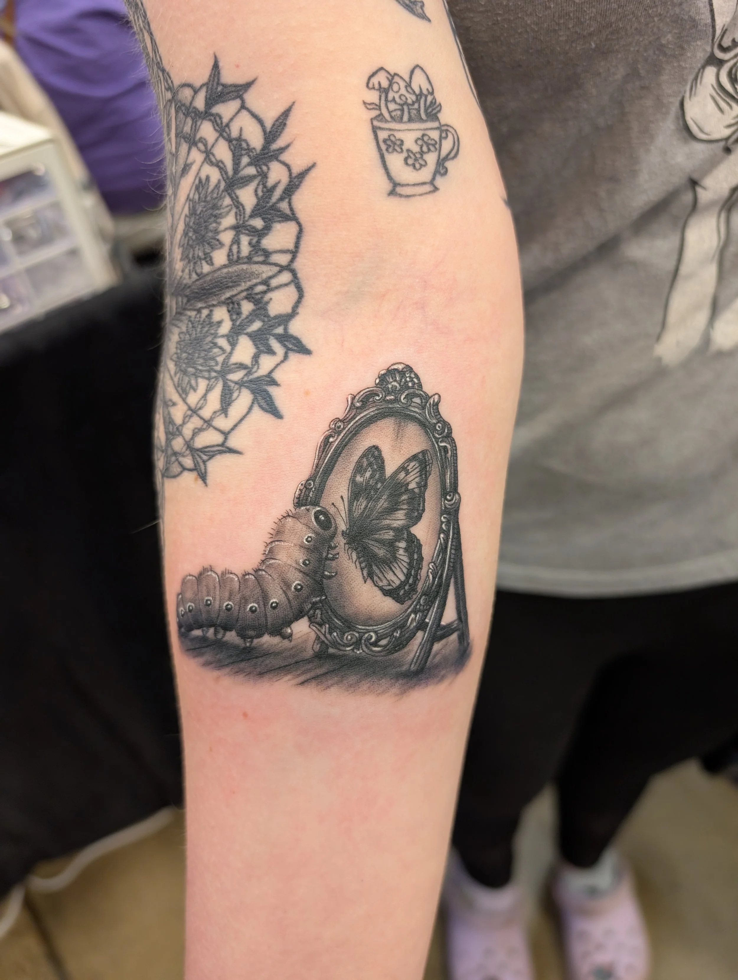 Tattoo of a butterfly inside a decorative mirror on a person's arm.