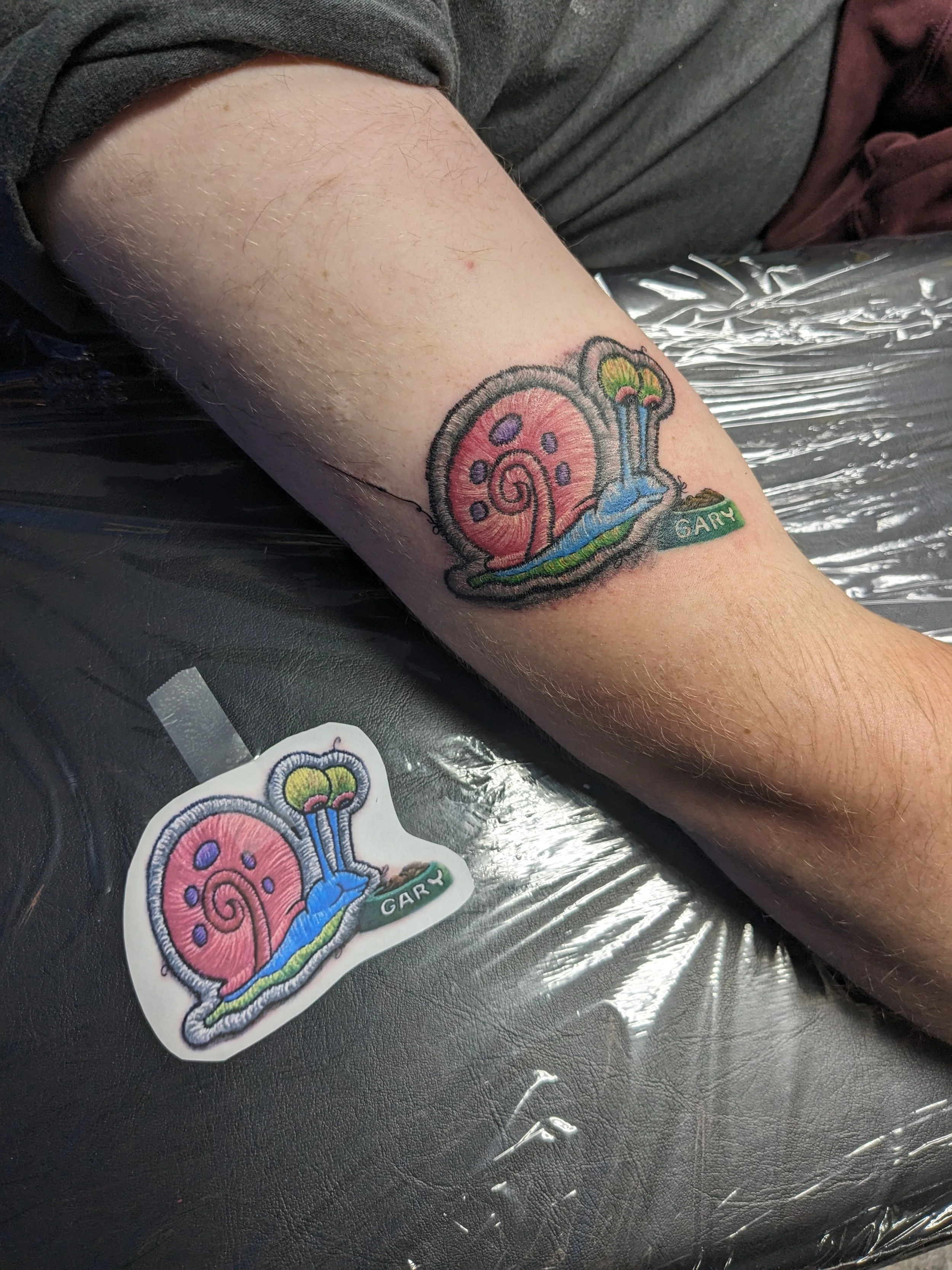 Close-up of a tattoo of a pink snail with purple spots and a yellow shell, along with a blue and green waterfall, and a green banner with the name "GARY" on a person's arm.