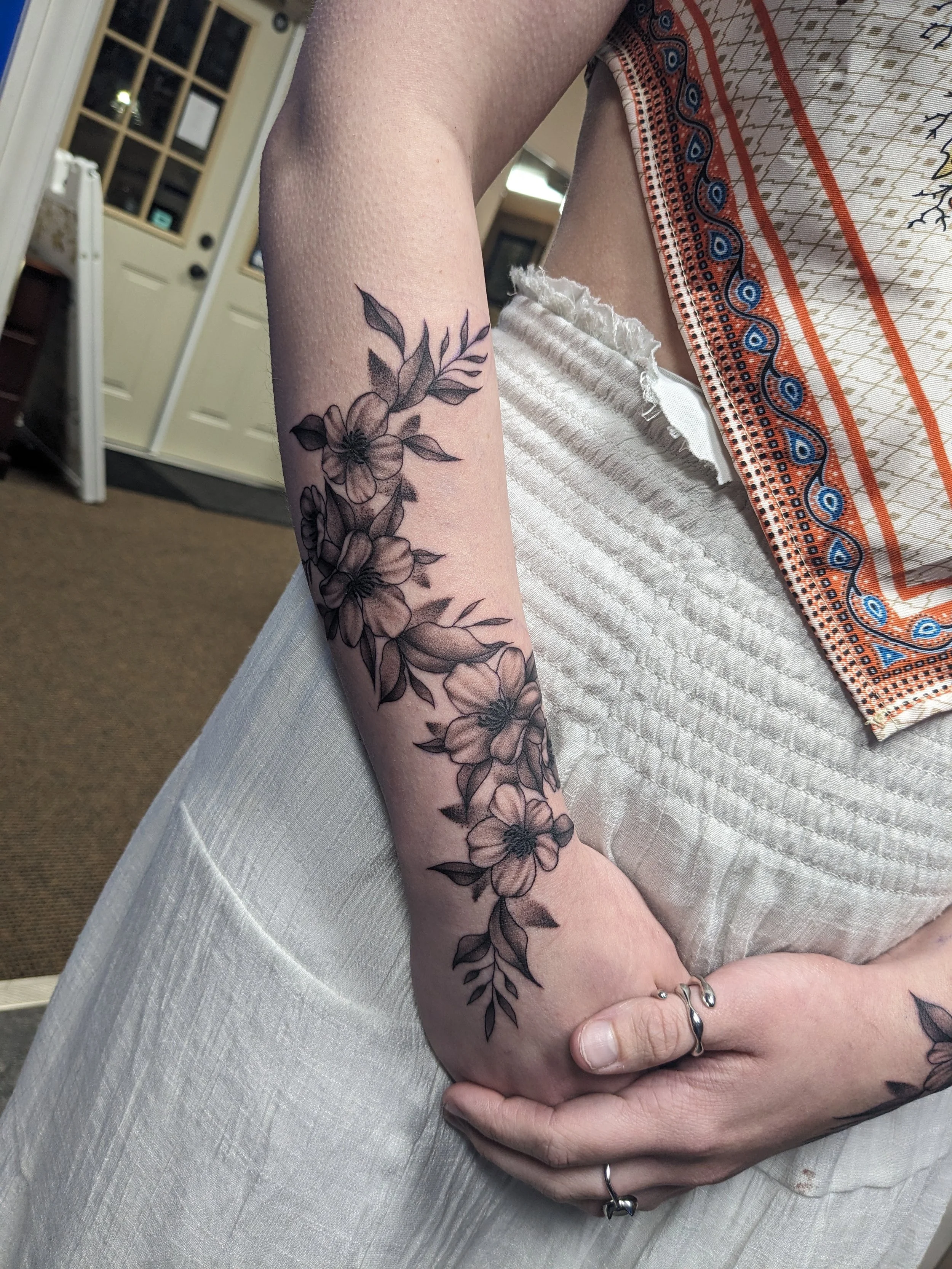 A person's arm with a floral tattoo, resting on their lap, wearing rings, sitting in front of a table with a patterned cloth. In the background, a door and cabinets are visible.