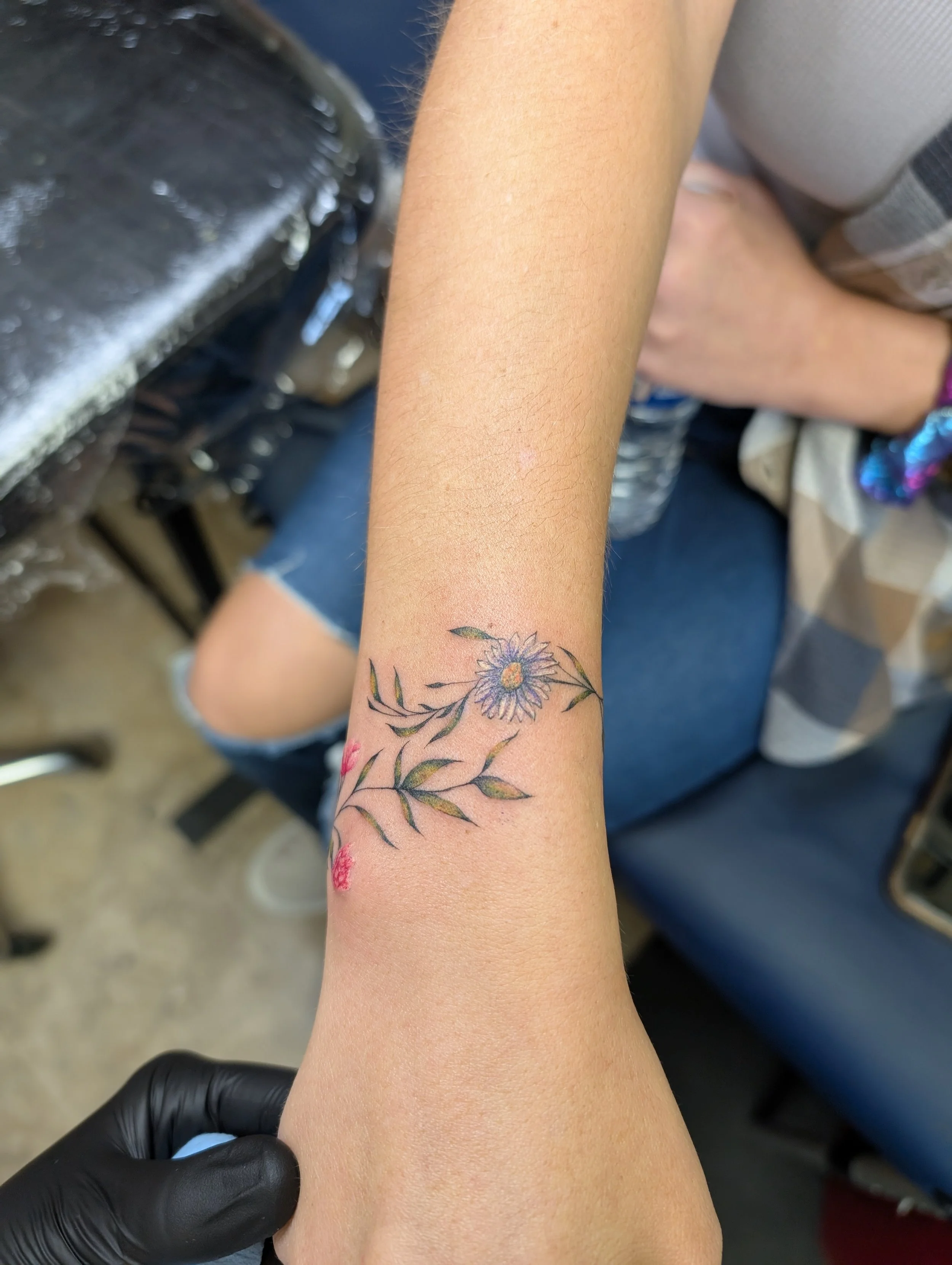 A person's arm with a colorful floral tattoo, held by a person wearing a black glove, with a background including a chair and water bottles.
