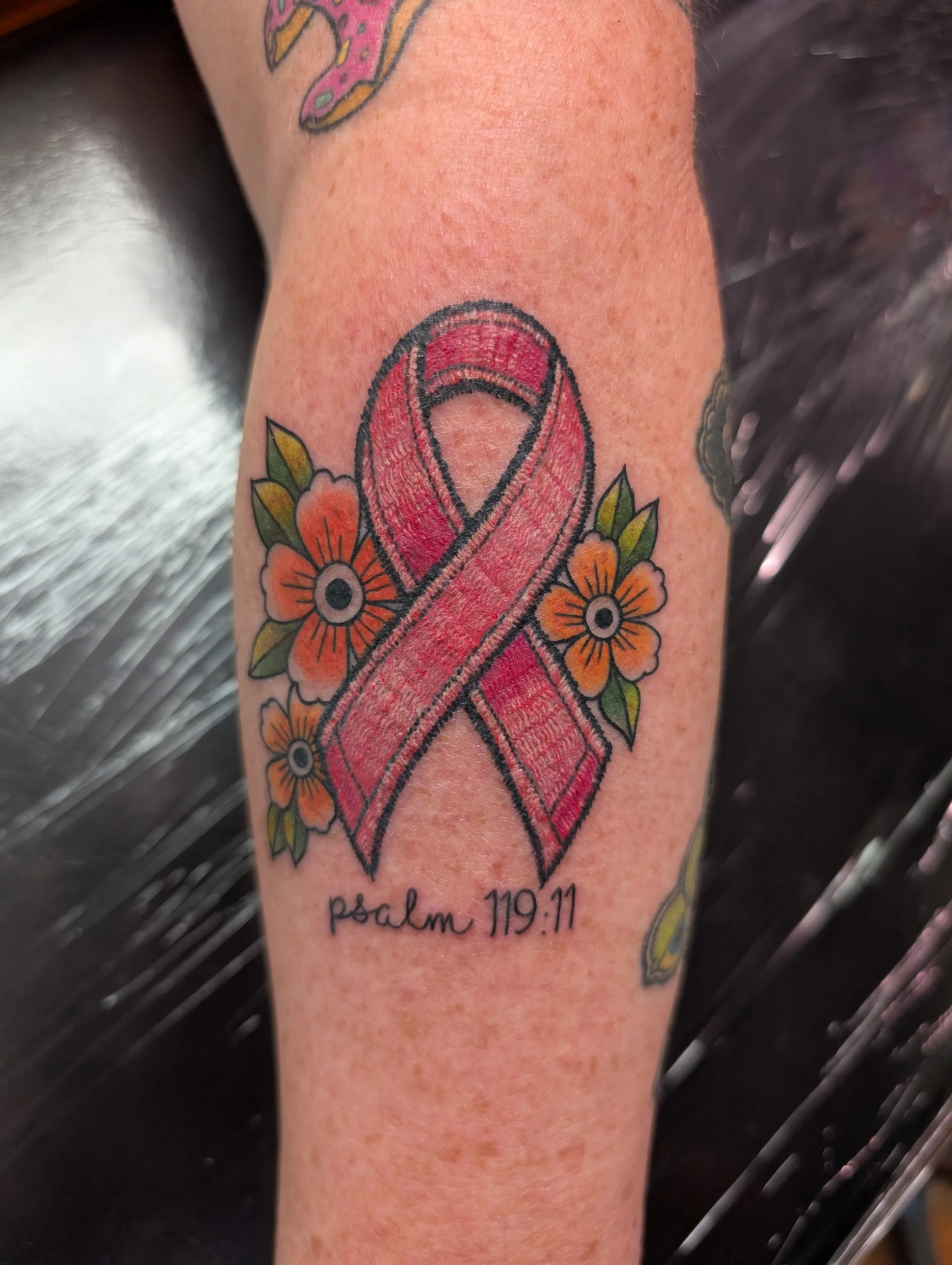Tattoo of a pink awareness ribbon surrounded by orange flowers with black centers on a person's forearm, with the words "psalm 119:11" written below.