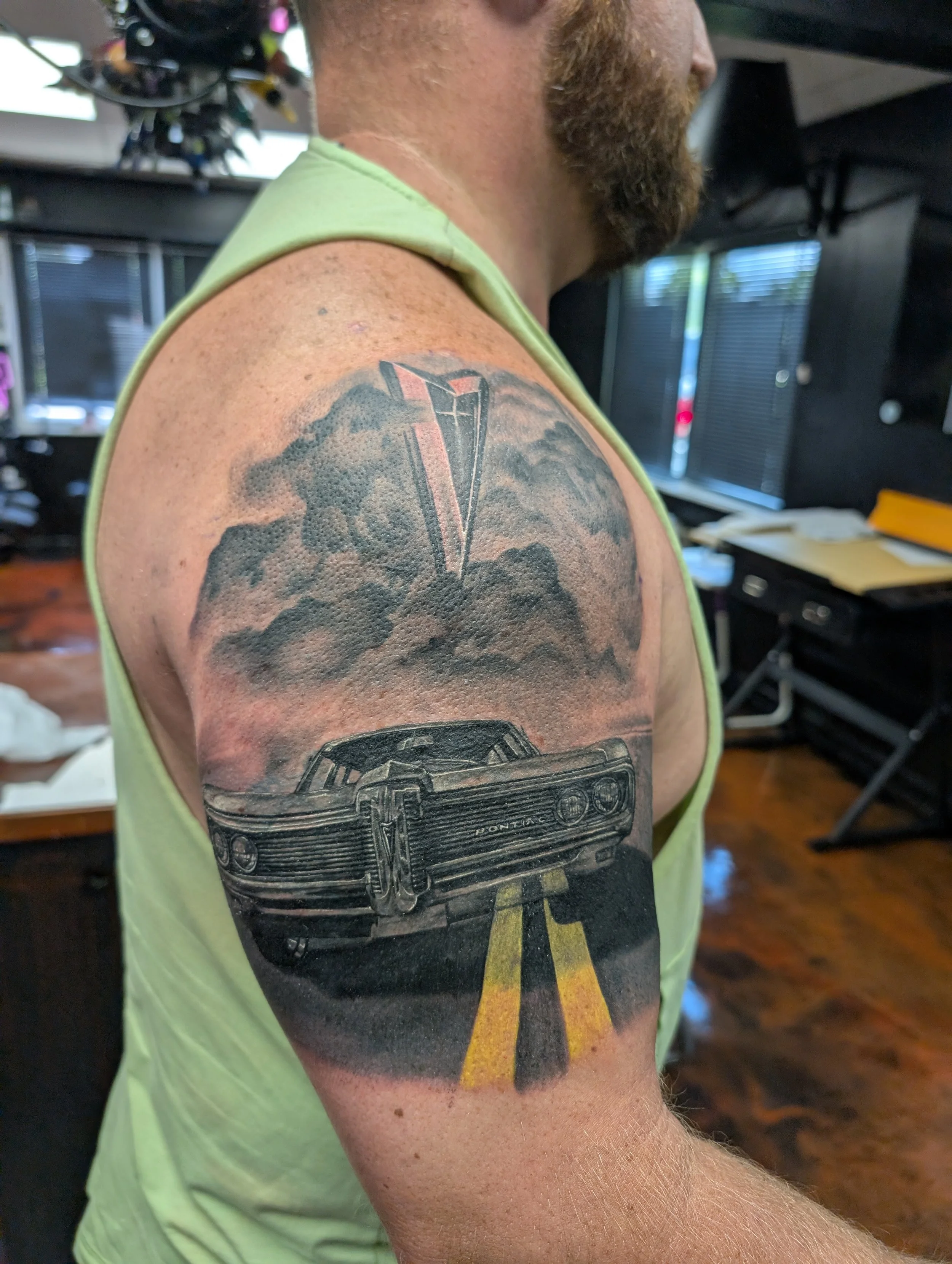 Close-up of a man's arm with a tattoo of a car and a background of clouds and a sunset sky, wearing a yellow sleeveless shirt, indoors with blinds and equipment in the background.