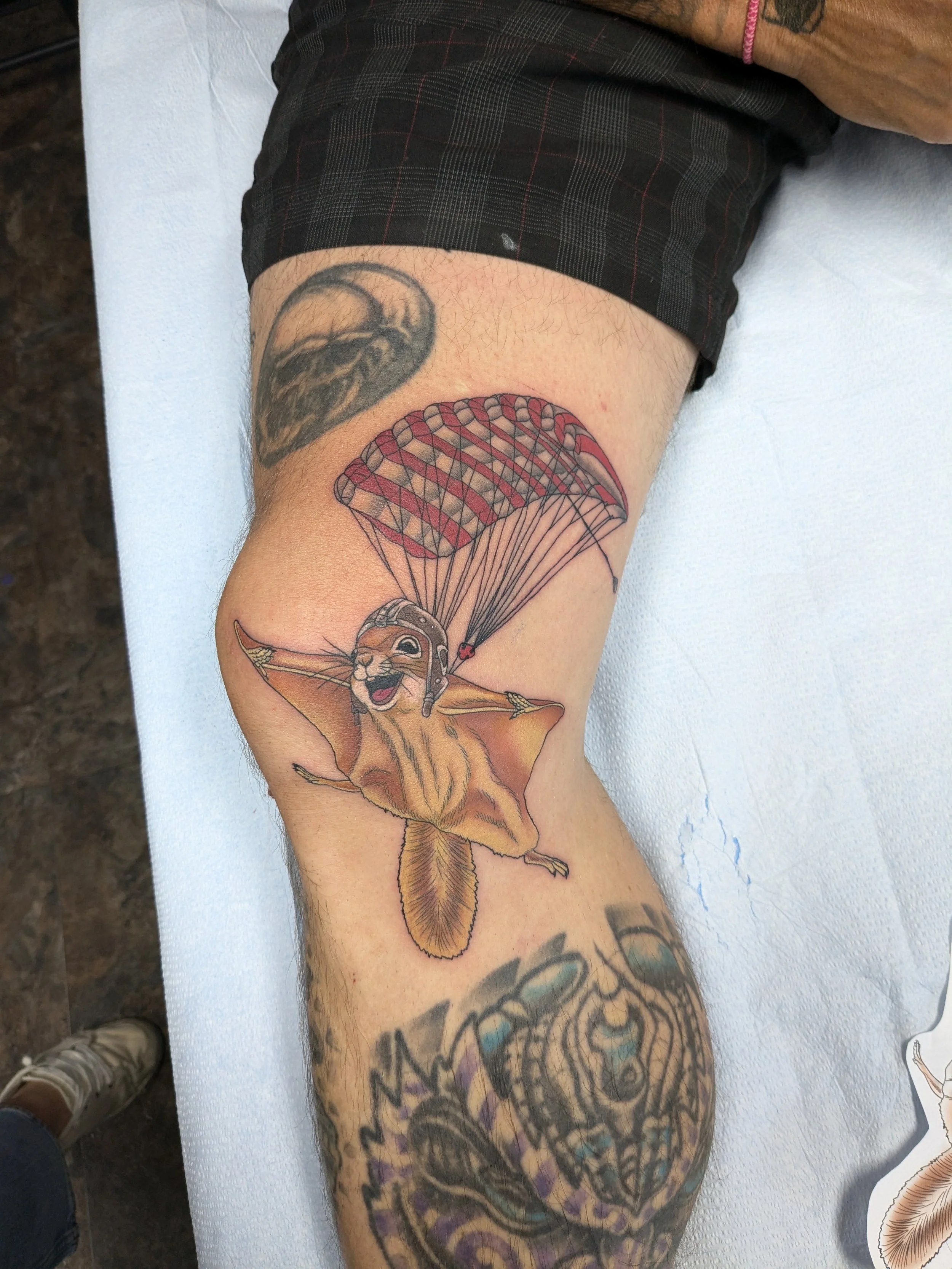 A person's arm with two tattoos: one of a moon with a tree inside, and another of a winged lion with a parachute.