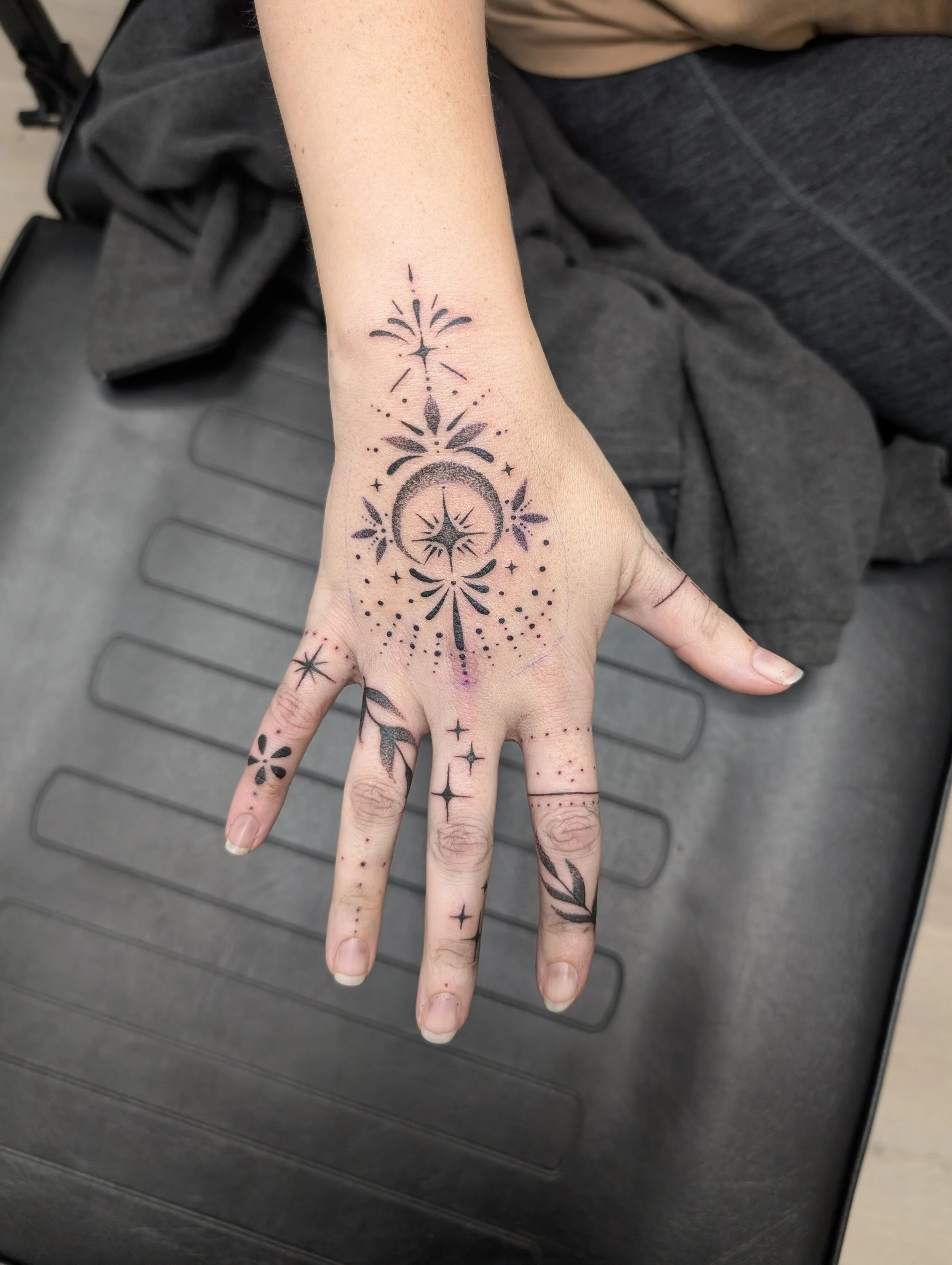 A person's hand with black tattoo designs, featuring stars, dots, floral shapes, and geometric elements, on the fingers, hand, and wrist, resting on a gray surface.