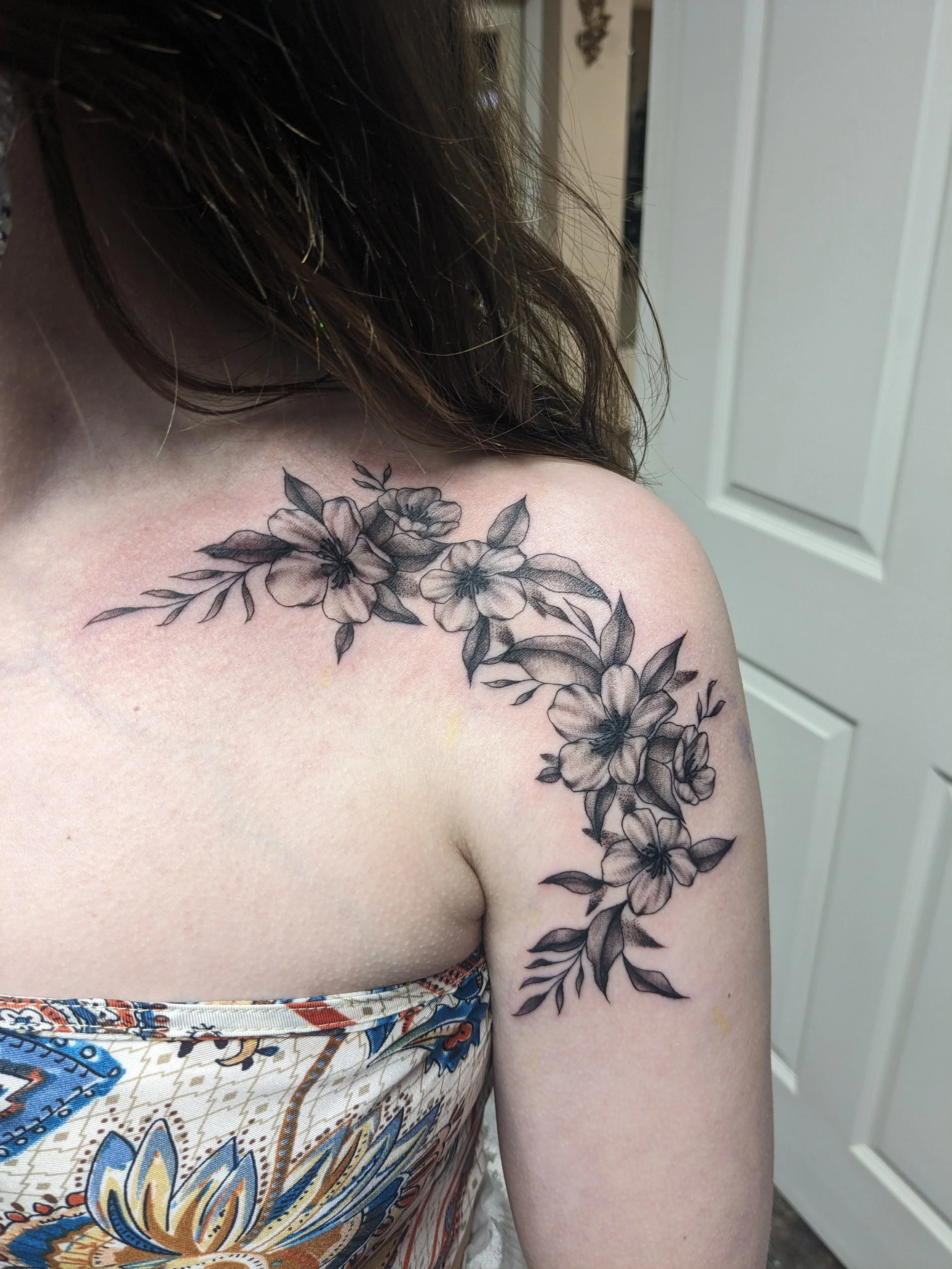 A close-up of a woman's shoulder and upper arm with a detailed black and gray floral tattoo, showing flowers and leaves, and part of her hair and patterned top are visible.