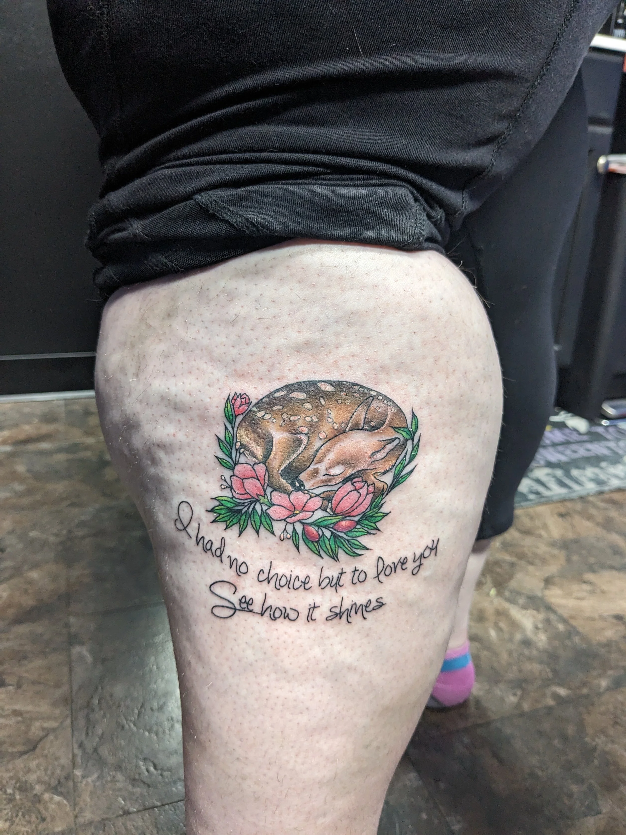 Tattoo of a fox curled up inside a circle of pink flowers and green leaves on a person's thigh, with a handwritten quote.