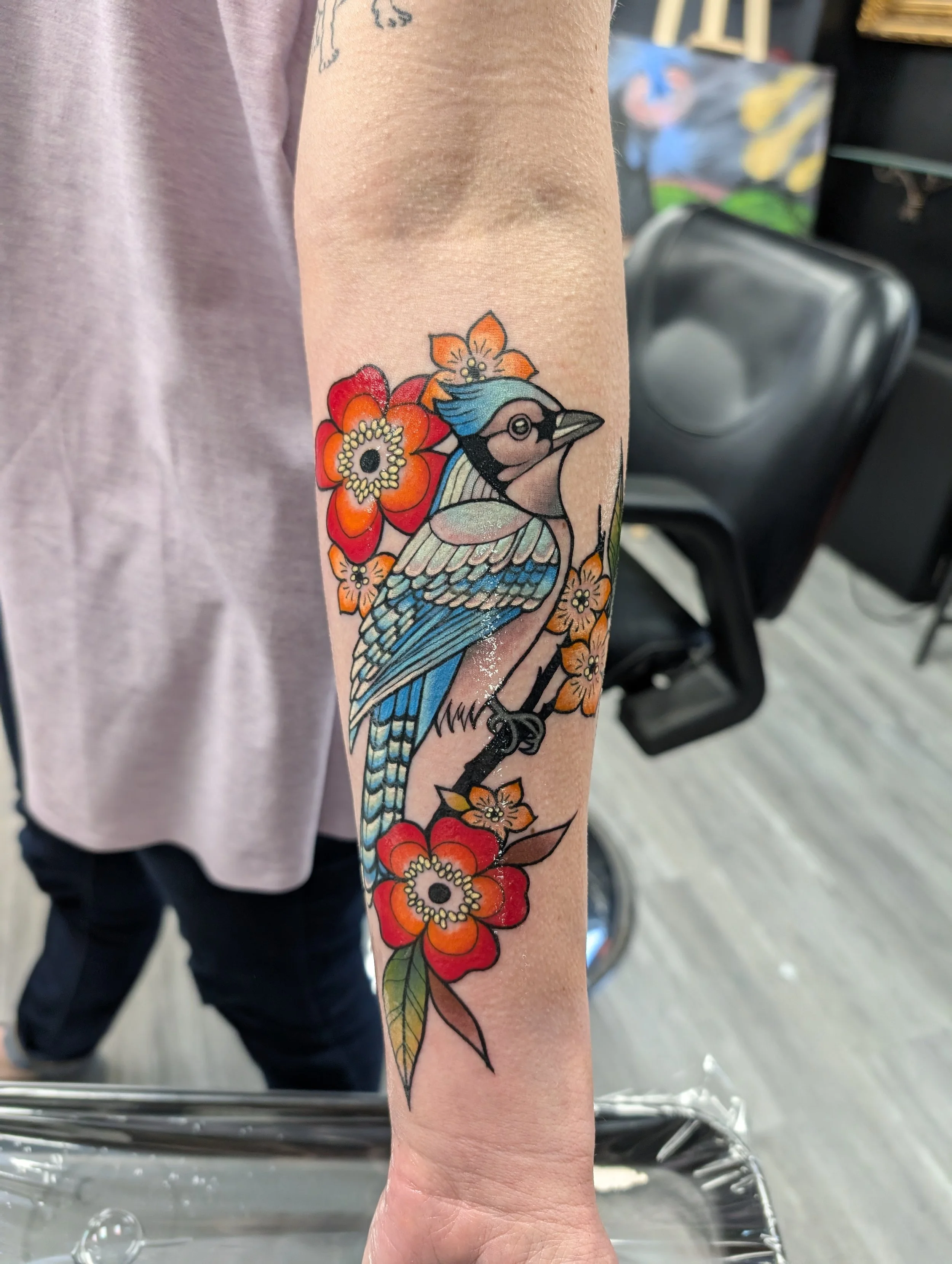 A colorful tattoo of a bird with blue, black, and beige feathers surrounded by orange and red flowers on a person's forearm.
