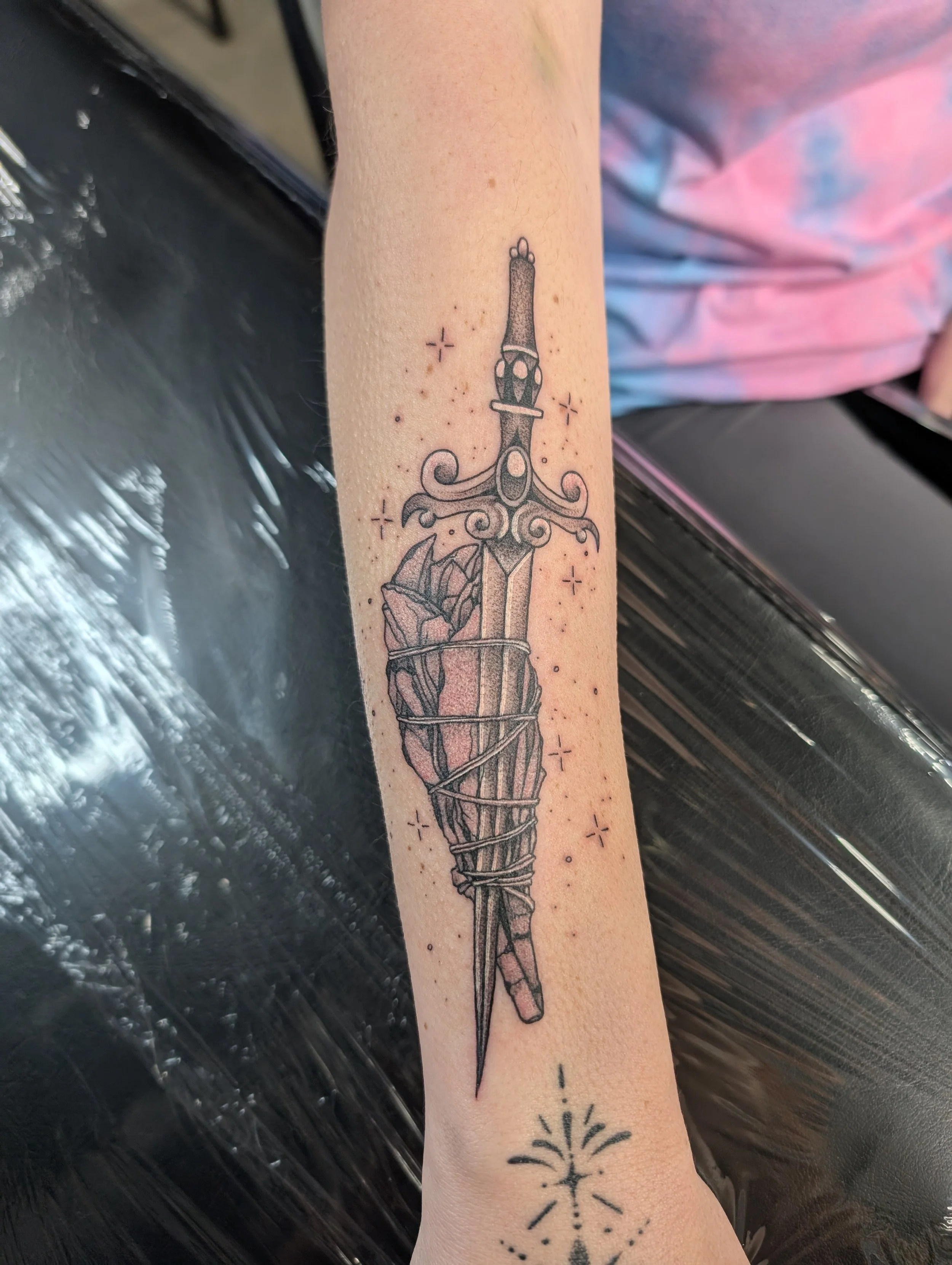 Black ink outline tattoo of a sword with a decorative handle, wrapped in a bandage or ribbon, with small star and dot accents around it, on a person's forearm.
