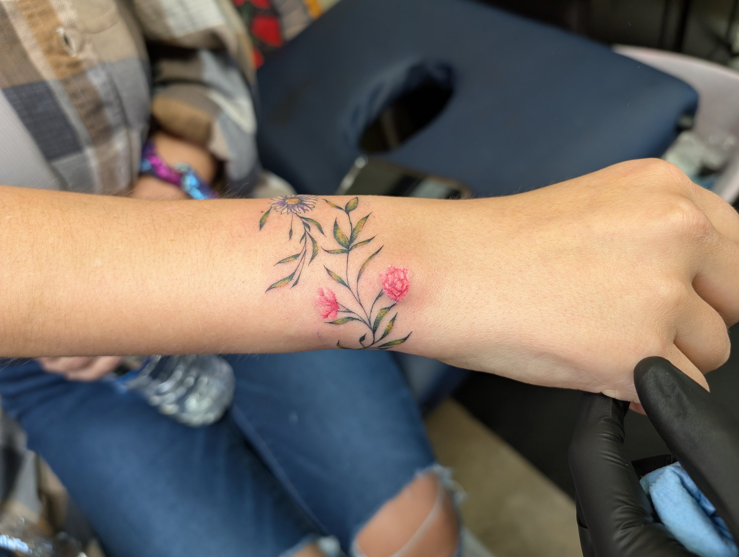 Close-up of a person's forearm with a colorful floral tattoo, holding a black object, with a blurred background showing casual clothing and indoor setting.