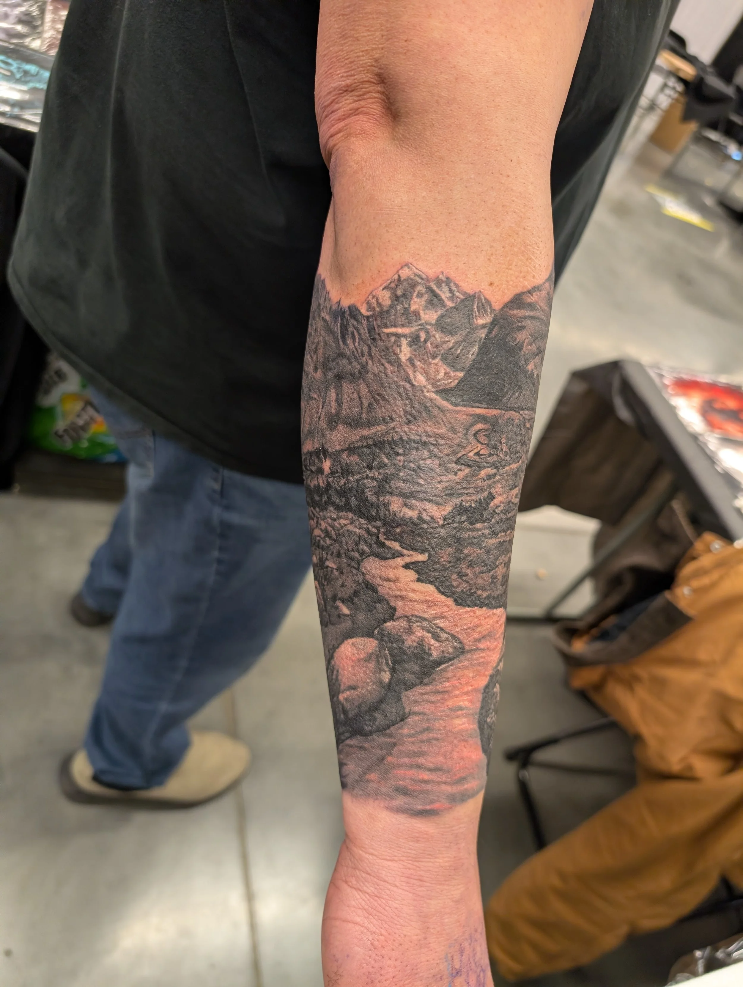 A close-up of a person's arm with a detailed black and gray mountain landscape tattoo, showing snowy peaks, rocky cliffs, and rugged terrain.