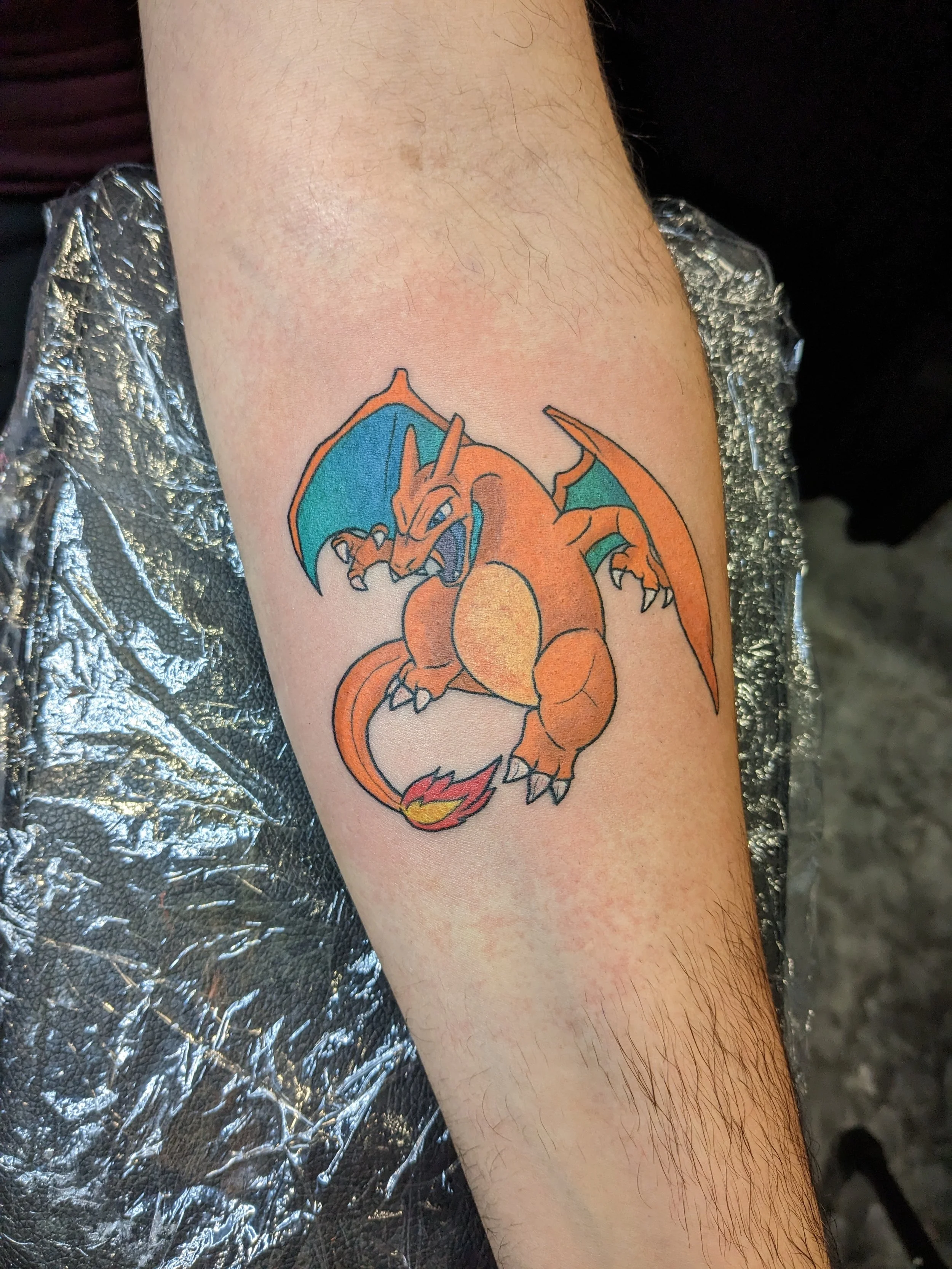 Colorful tattoo of Charizard, a dragon-like Pokémon, on a person’s upper arm with a background of metallic silver material.