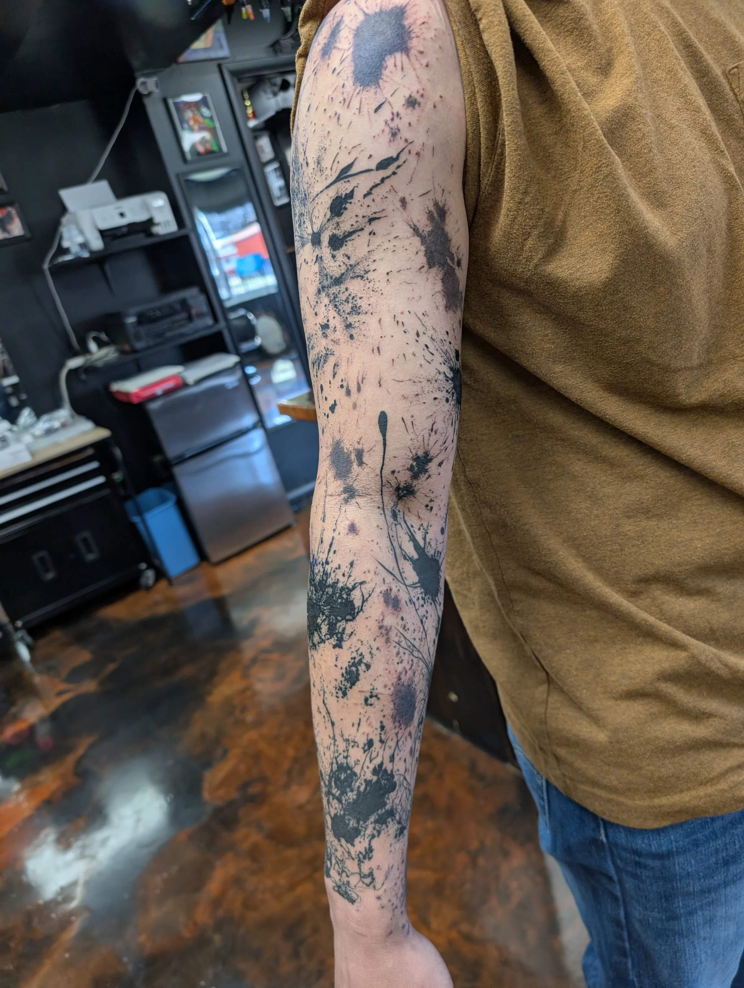 Tattooed arm with black ink splatters and abstract designs, wearing a tan sleeve shirt, in a room with black shelves and a computer setup in the background.