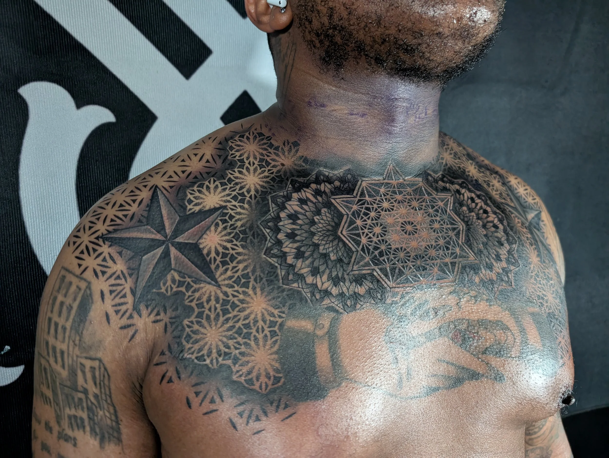 Close-up of a person's upper chest, neck, and face showing intricate geometric tattoos covering the chest and shoulder, with some facial hair visible.