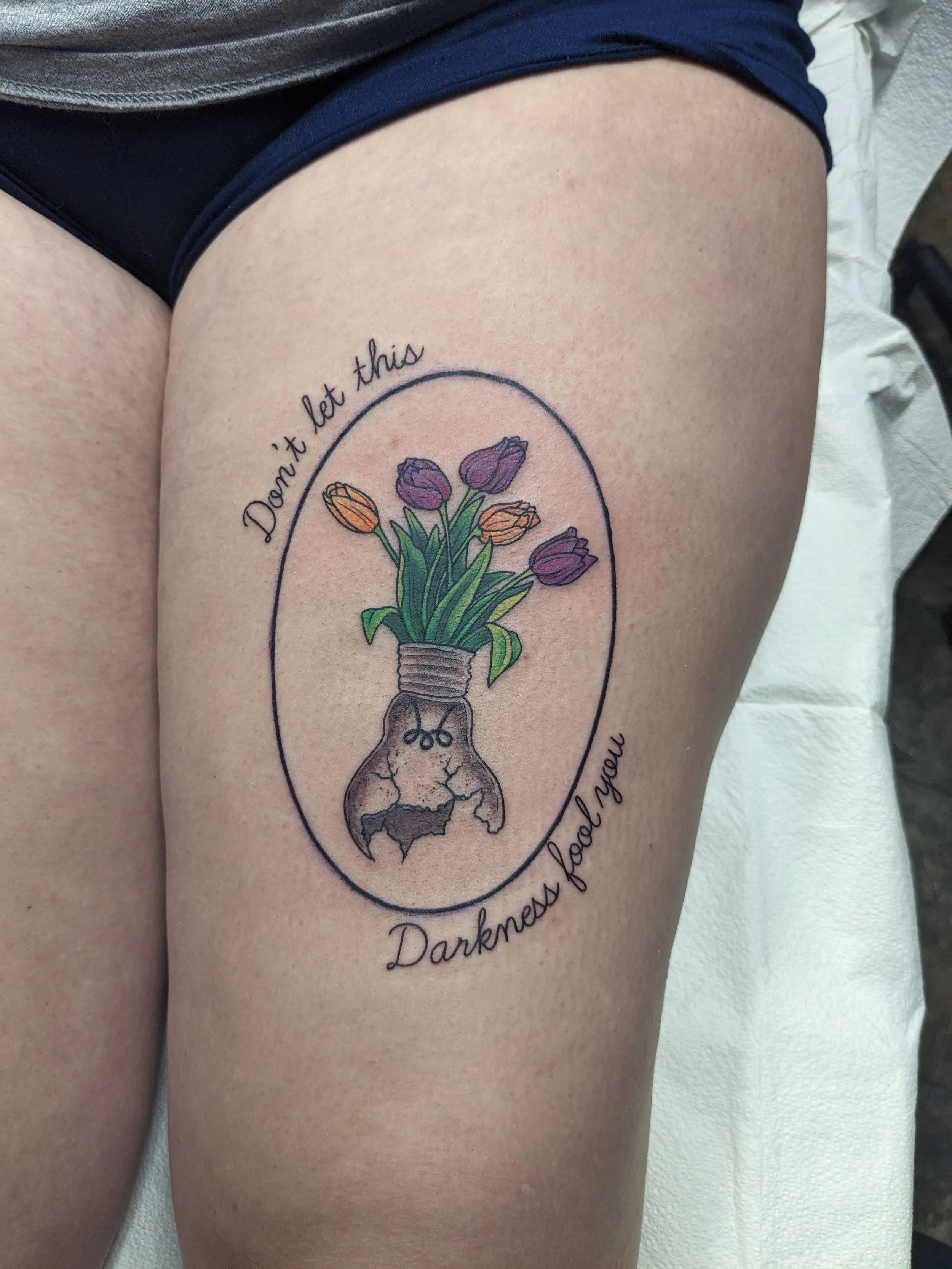 Tattoo of a light bulb with colorful flowers inside, along with the phrase 'Darker days don't let this darkness' written in cursive.
