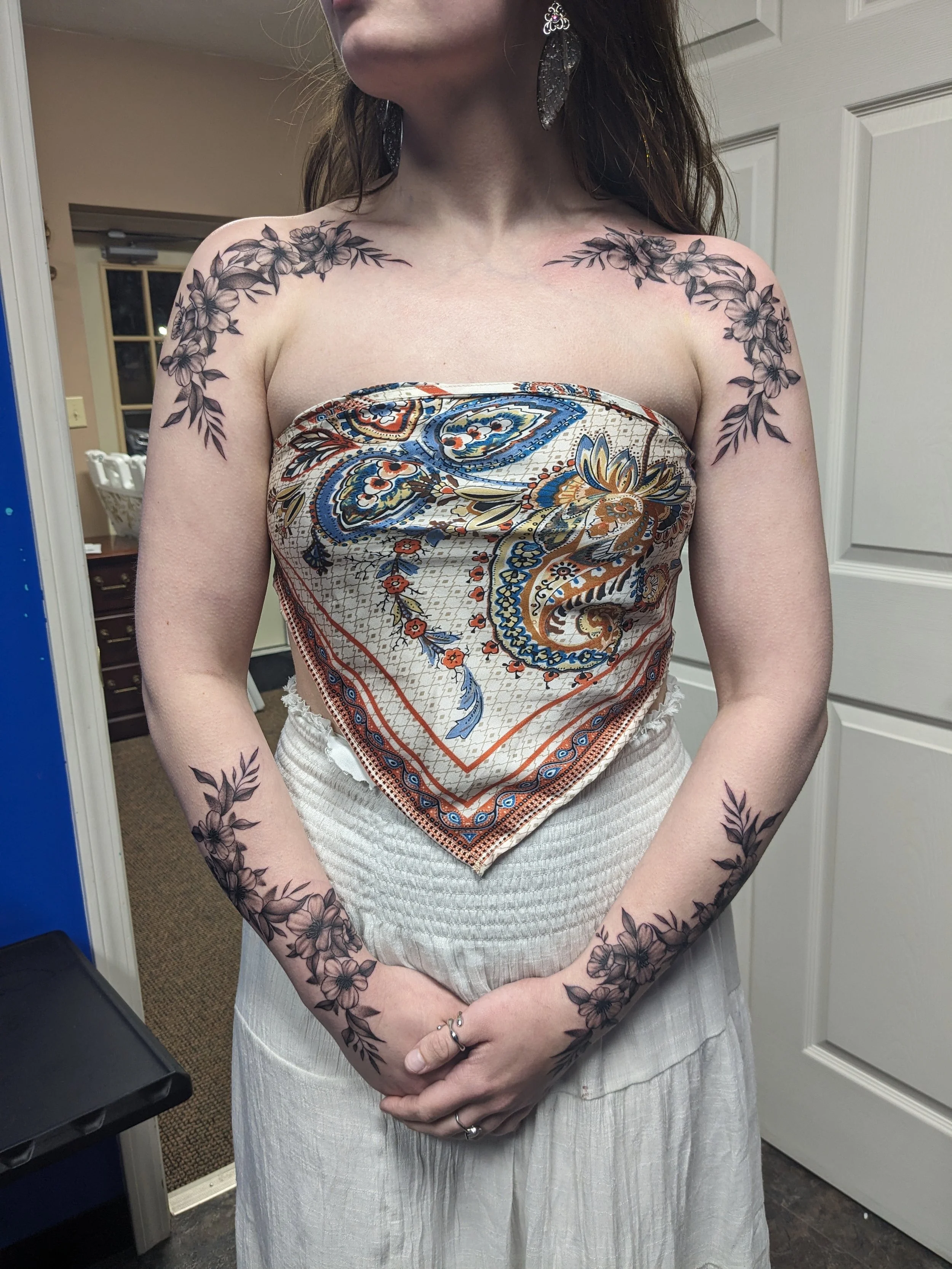 A woman with floral tattoos on her arms, shoulders, and hands, wearing a patterned tube top and a white skirt, standing indoors near a mirror.