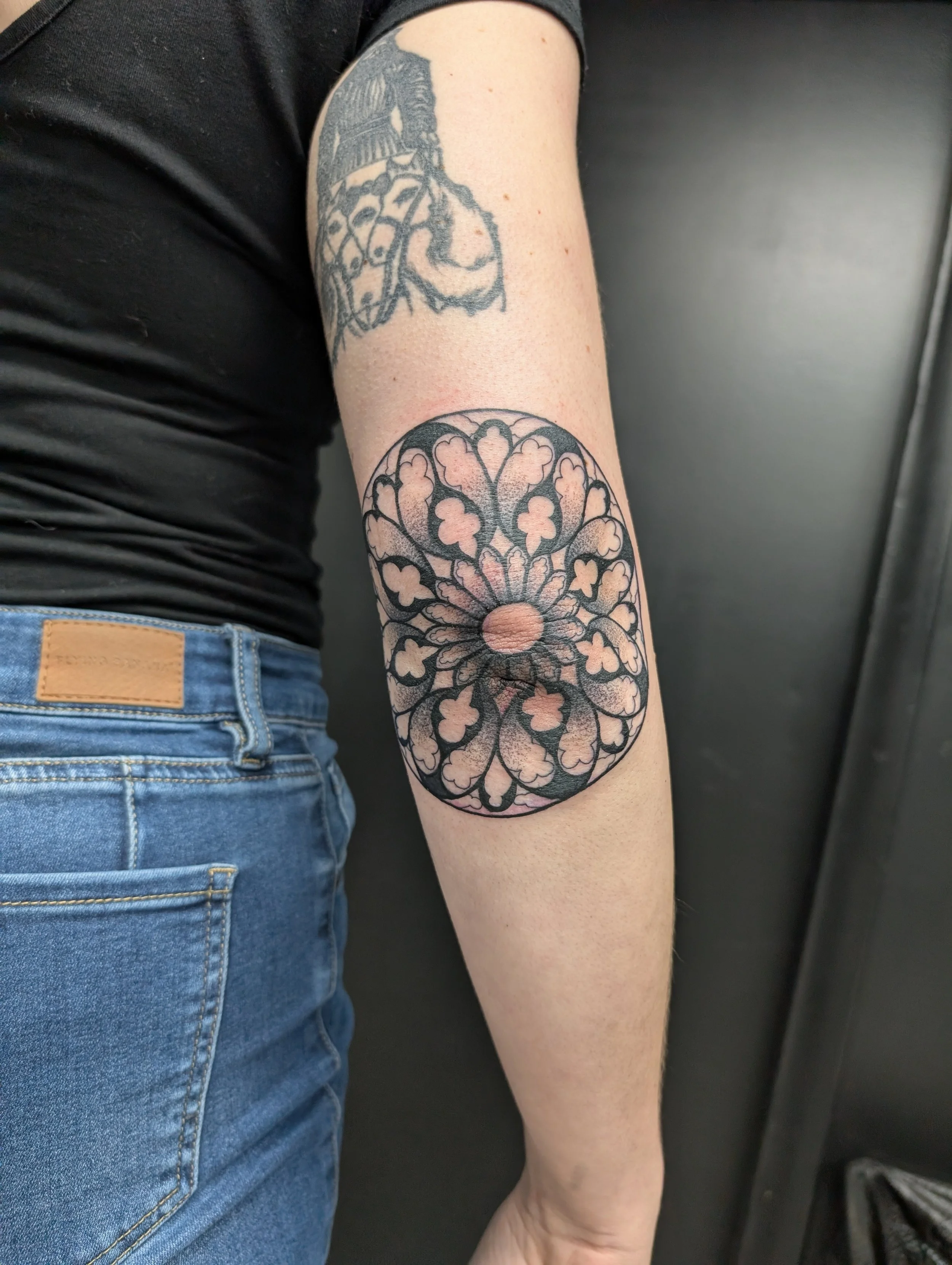A person's arm with two tattoos, one of a jester playing cards and the other a flower mandala design, next to a pair of blue jeans and a black shirt.