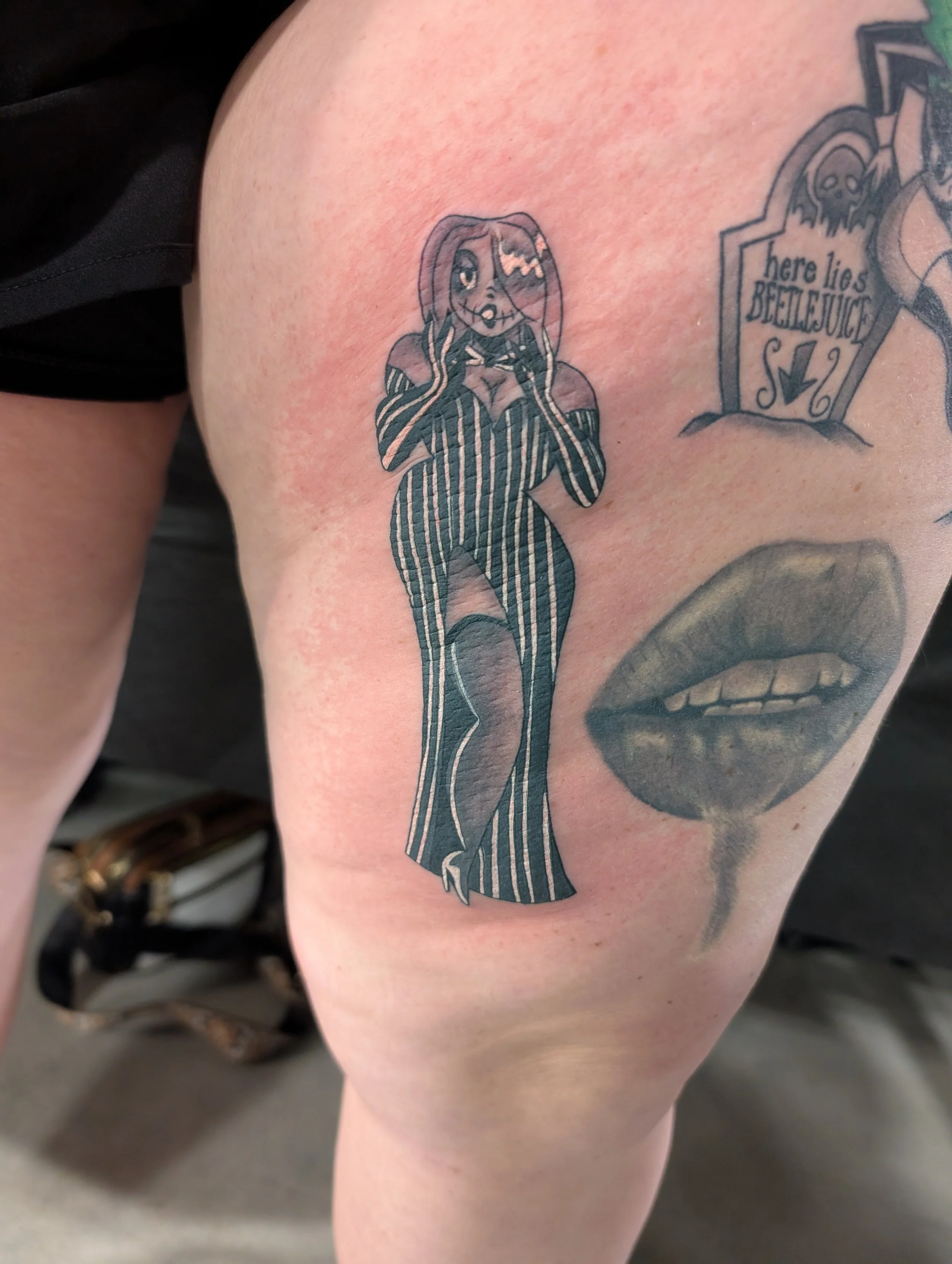 Tattoo of a cartoon woman with long hair, one eye closed, making a wink, wearing a dress, holding her hands to her face, on person's thigh.