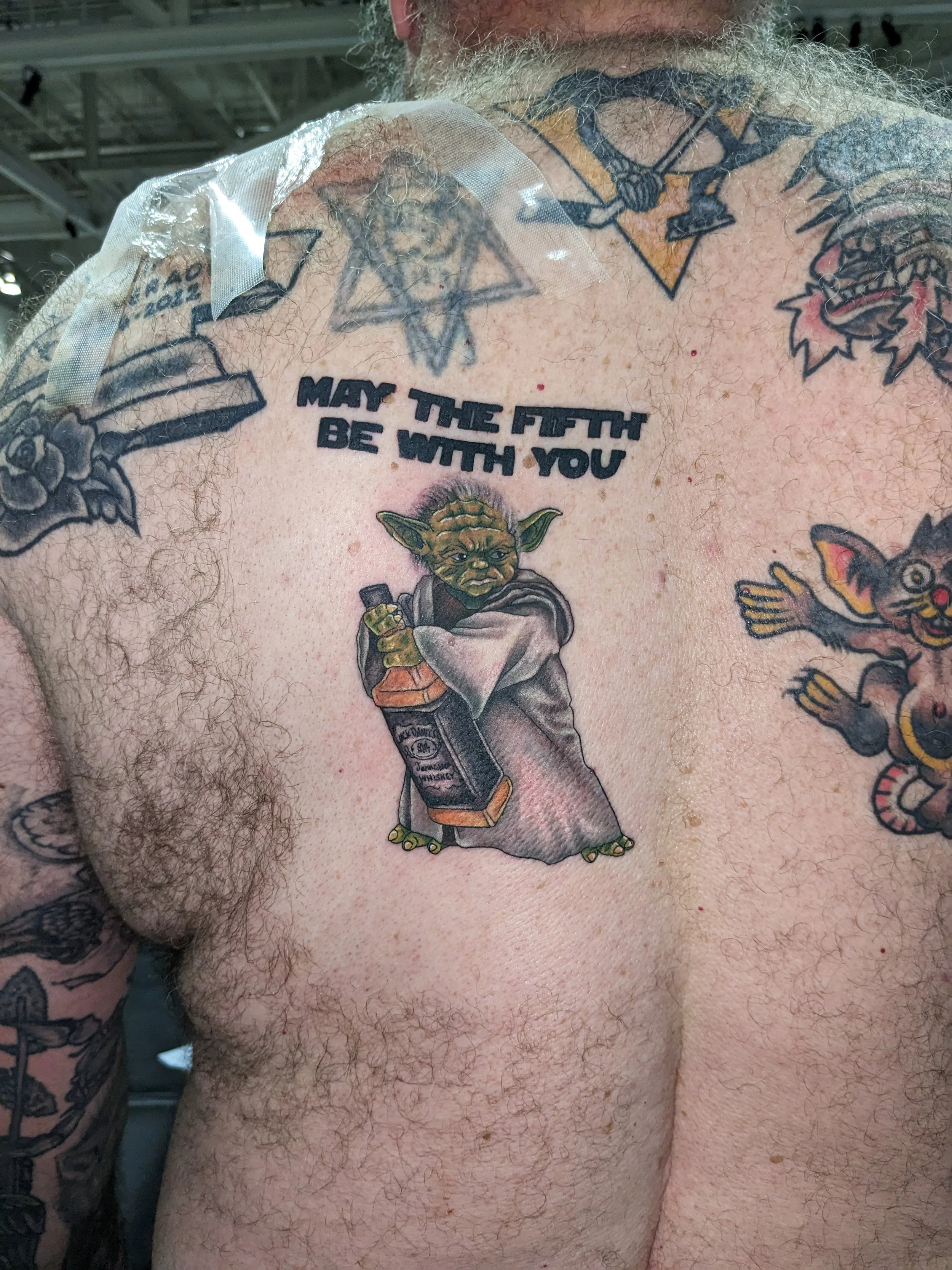 Close-up of a person's chest and shoulder with multiple tattoos, including a Jedi Yoda holding a beer bottle, a Star of David with a sword, and a phrase that reads "MAY THE FIFTH BE WITH YOU." The person has light skin, body hair, and some redness ar