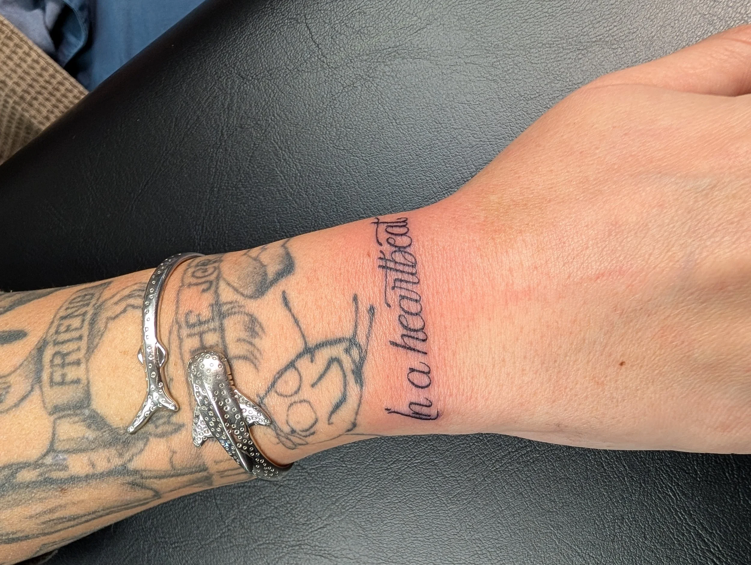 Close-up of a person's forearm tattoo with cursive writing, wearing multiple silver bracelets, resting on a black textured surface.