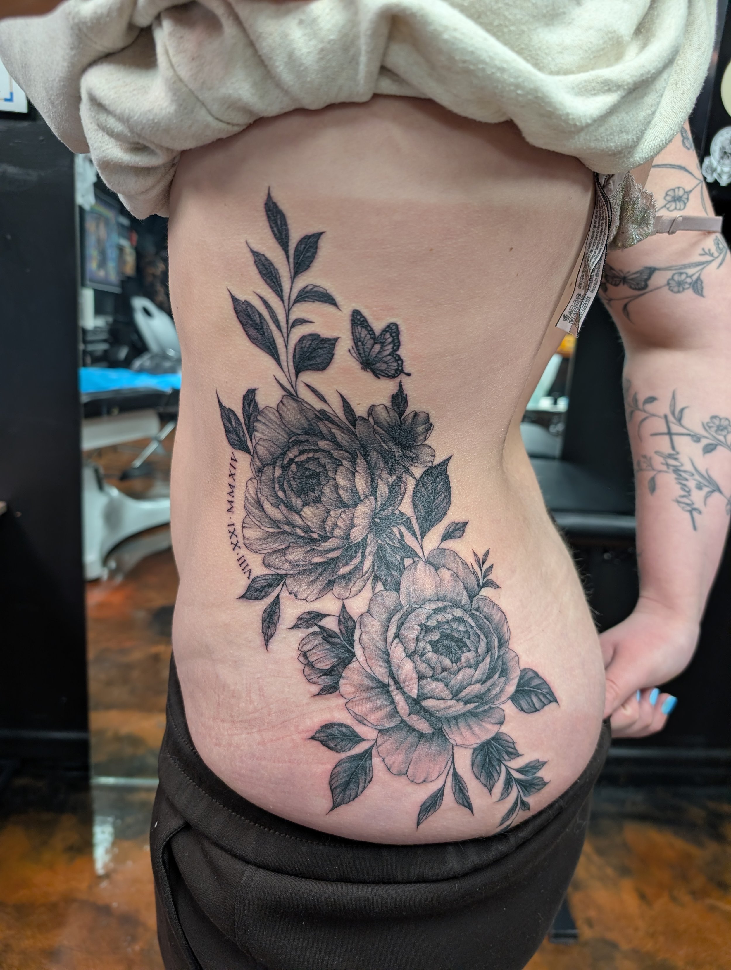 A person with a floral tattoo on their side and arm, with the tattoo featuring detailed flowers, leaves, and a butterfly. The person is wearing a beige shirt and black pants, standing indoors.