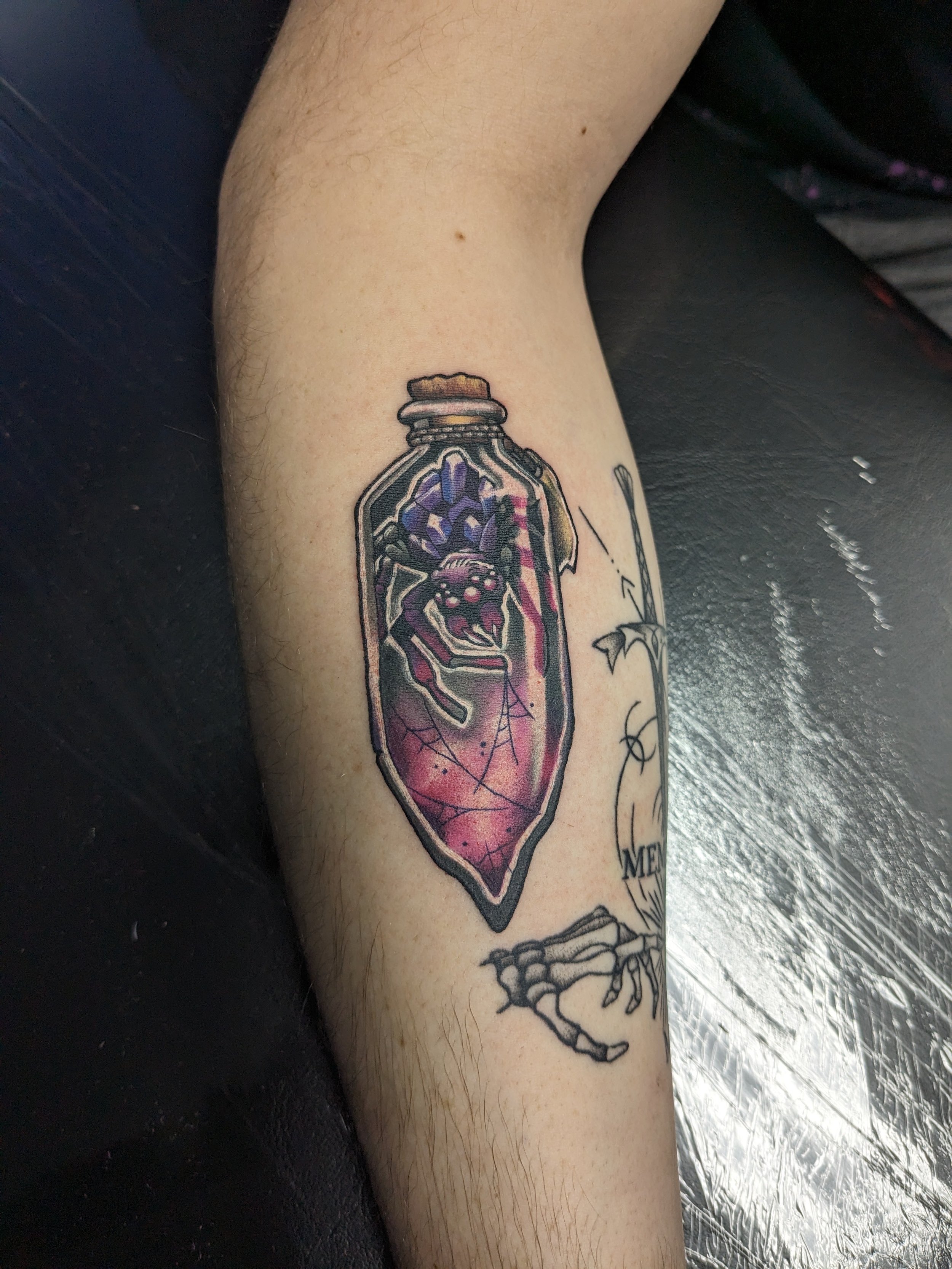 Tattoo of a vial containing purple and pink crystals on a person's arm with a skeletal hand and some text.