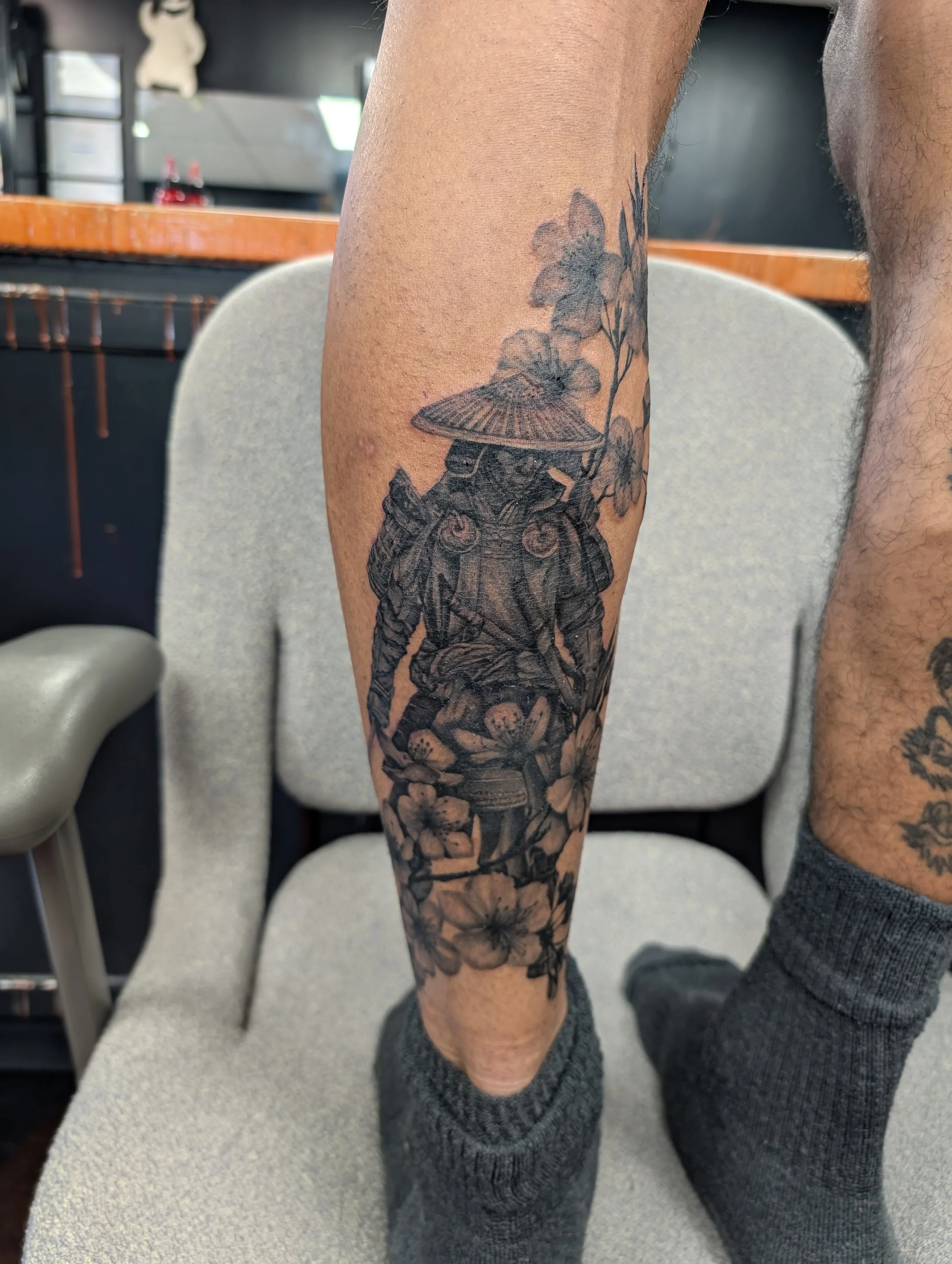 A person's forearm with a detailed black and gray tattoo of a drone surrounded by flowers, with a background of chairs and a table.