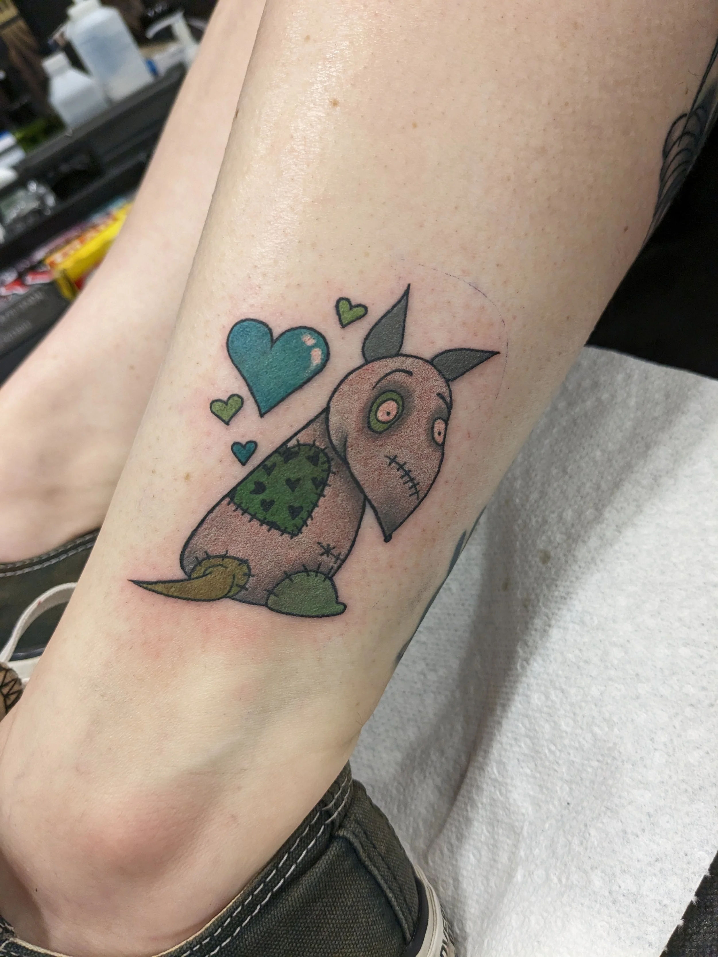 A colorful tattoo of cartoon-like creature with large round eyes, stitched mouth, green heart-shaped body, and a downward-pointing tail, surrounded by small green, blue, and teal hearts.