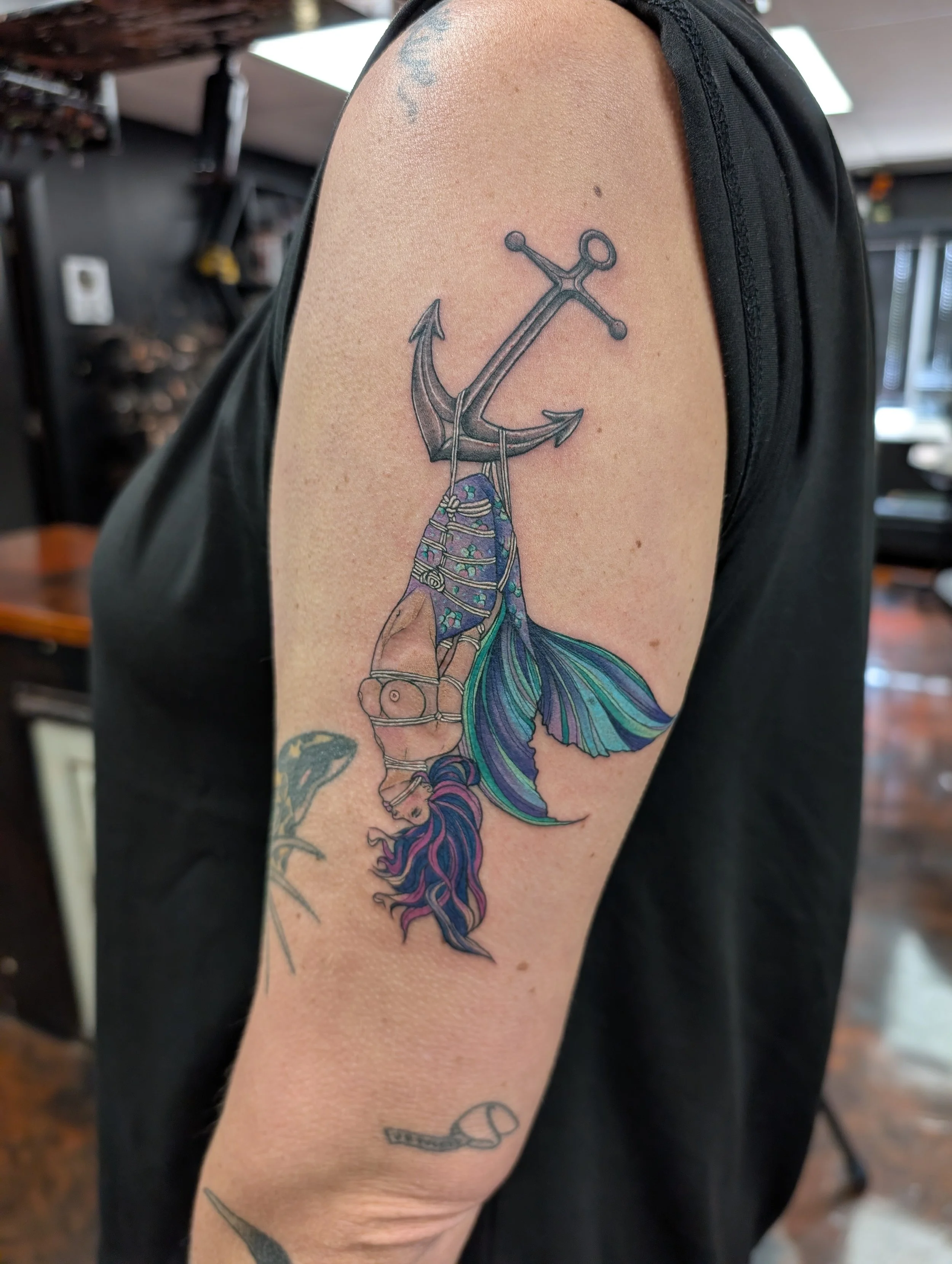 A tattoo of an anchor on a person's upper arm, with the anchor's chain forming a wave pattern. The tattoo has some colorful elements, including purple, blue, and green shades in the wave and chain area.