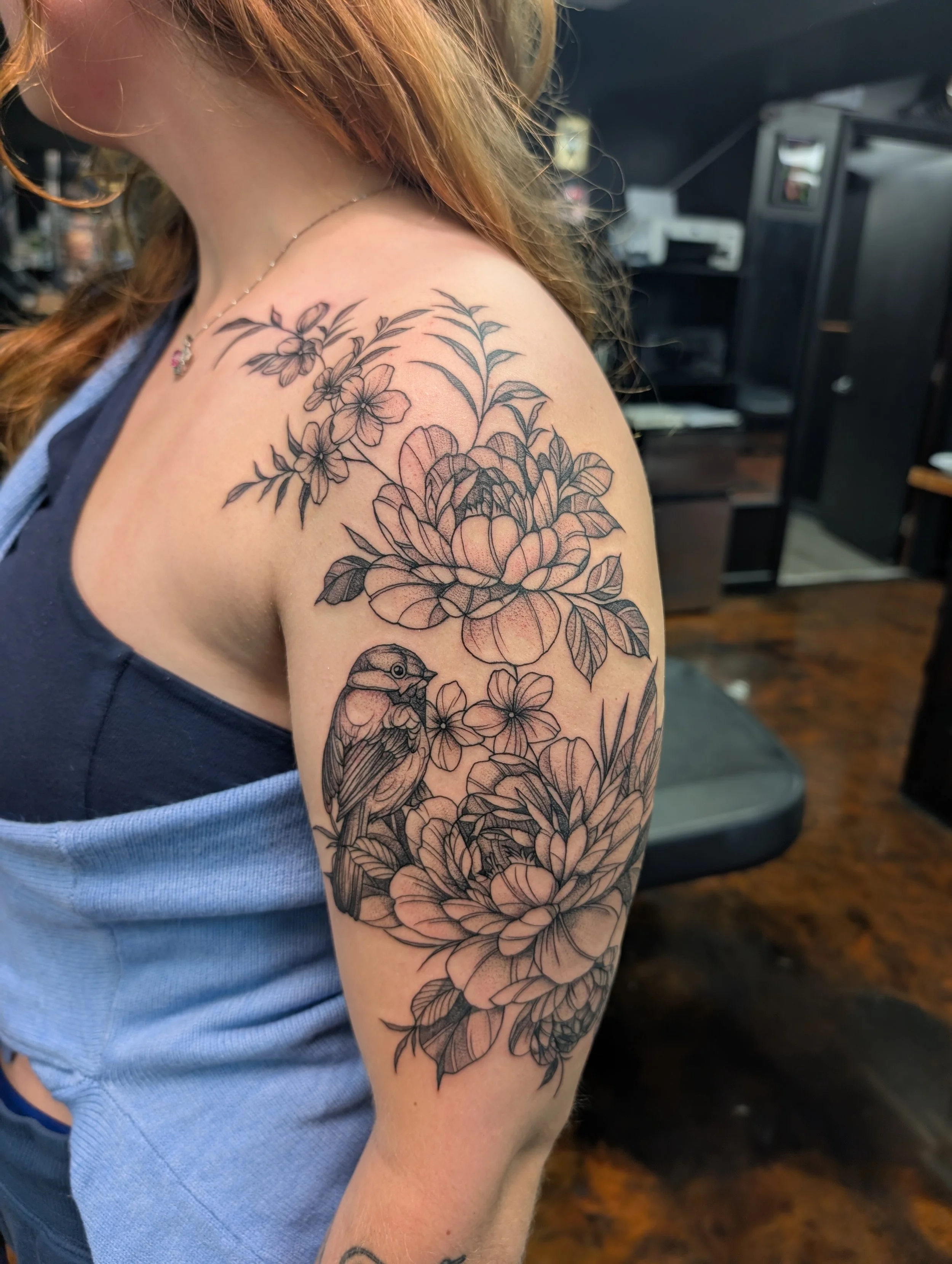 A person's shoulder and upper arm with a floral tattoo featuring large flowers, leaves, and a small bird.