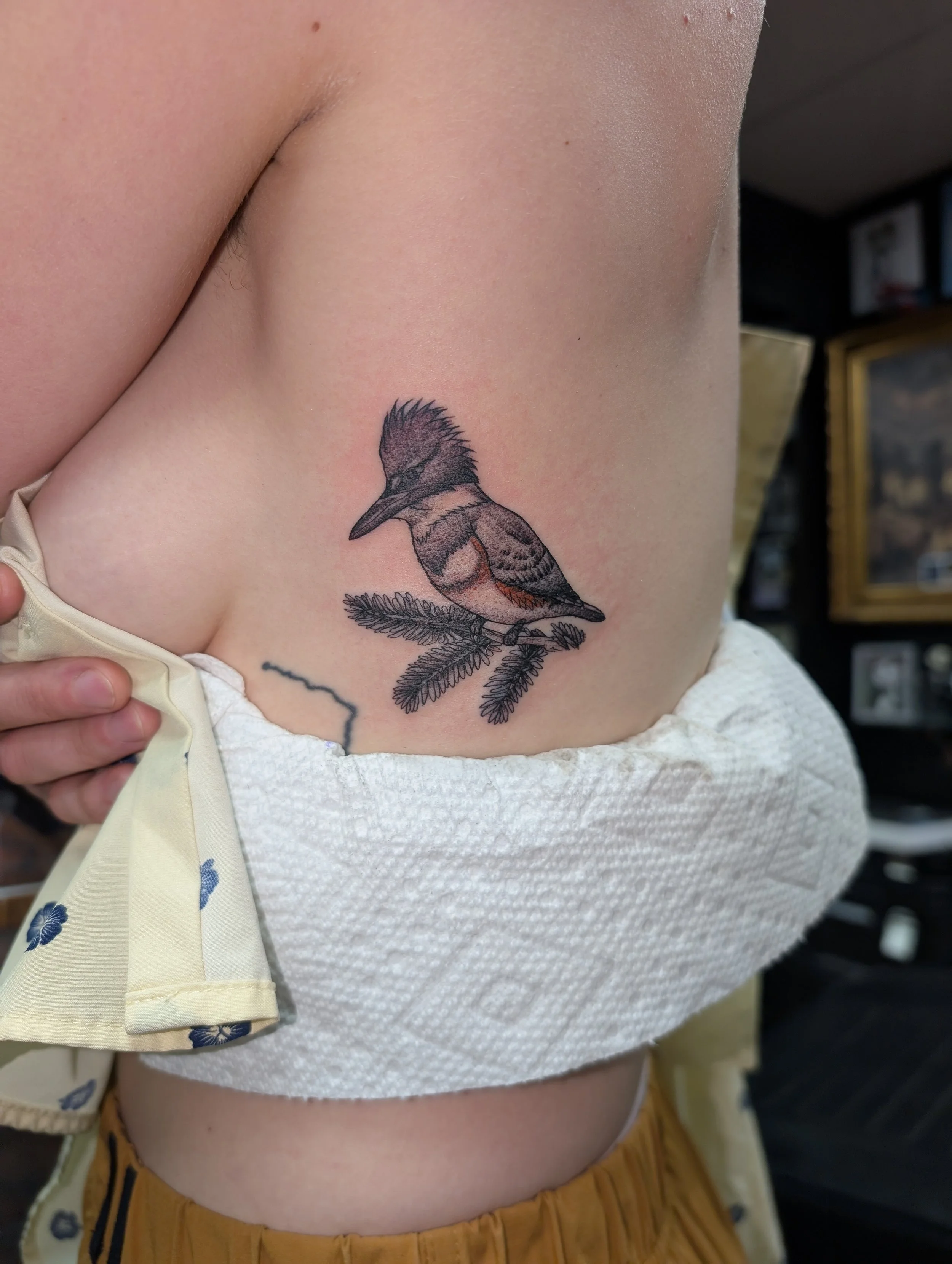 Close-up of a person's side showing a bird tattoo on their ribs, with part of their hand and beige clothing visible.