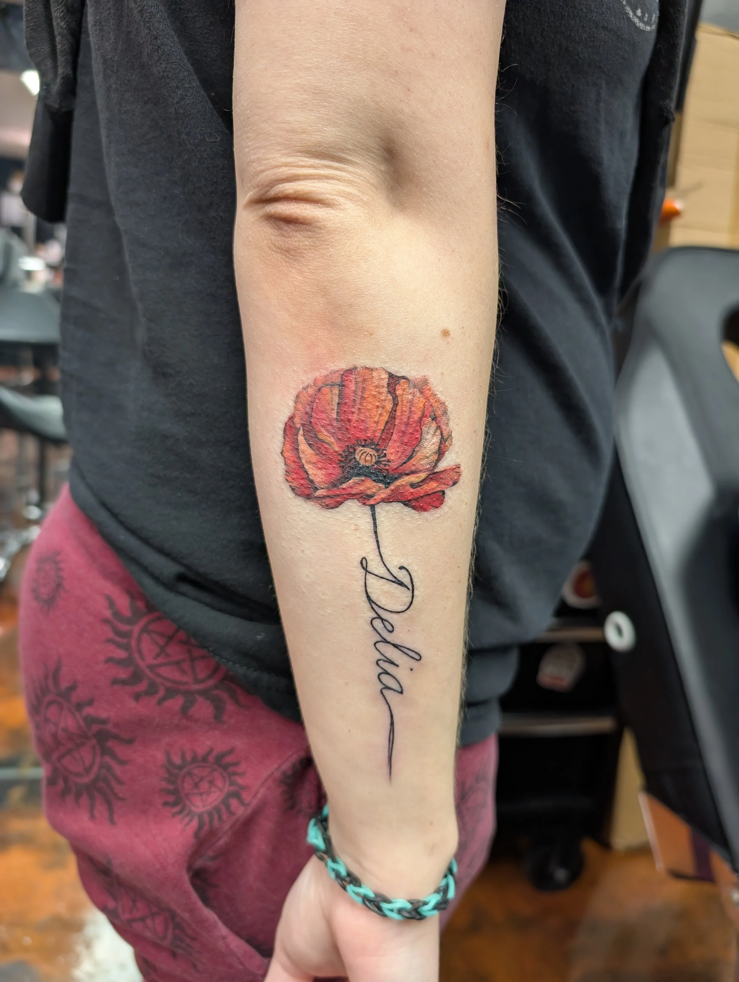 A person's right forearm with a red poppy flower tattoo and the word "Dawn" written in cursive below it. The person is wearing a black bracelet and a turquoise and black plastic chain bracelet. The background includes a pinkish red garment and part o