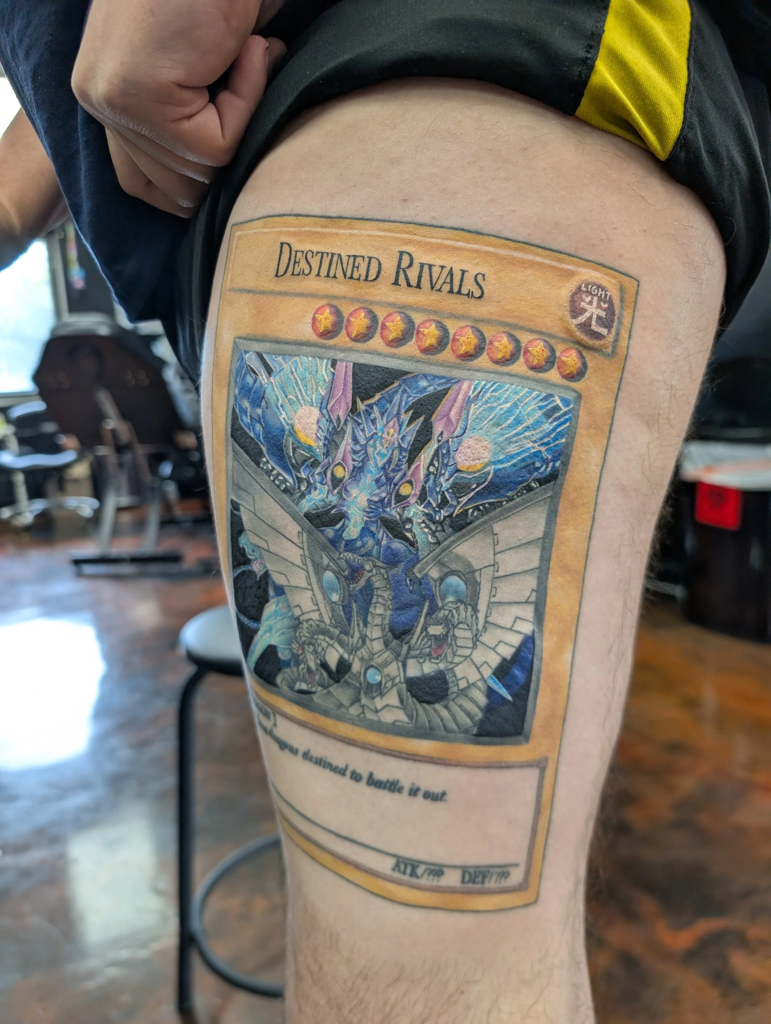 Tattoo on person's thigh featuring a card labeled 'Destined Rivals' with a dragon illustration and symbols, and text about battling it out.