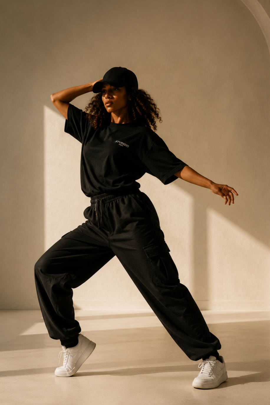 Femme noire en tenue de sport noire, cap, et baskets blanches, posant dans une pièce lumineuse avec un mur beige, en mouvement. Cannetille.