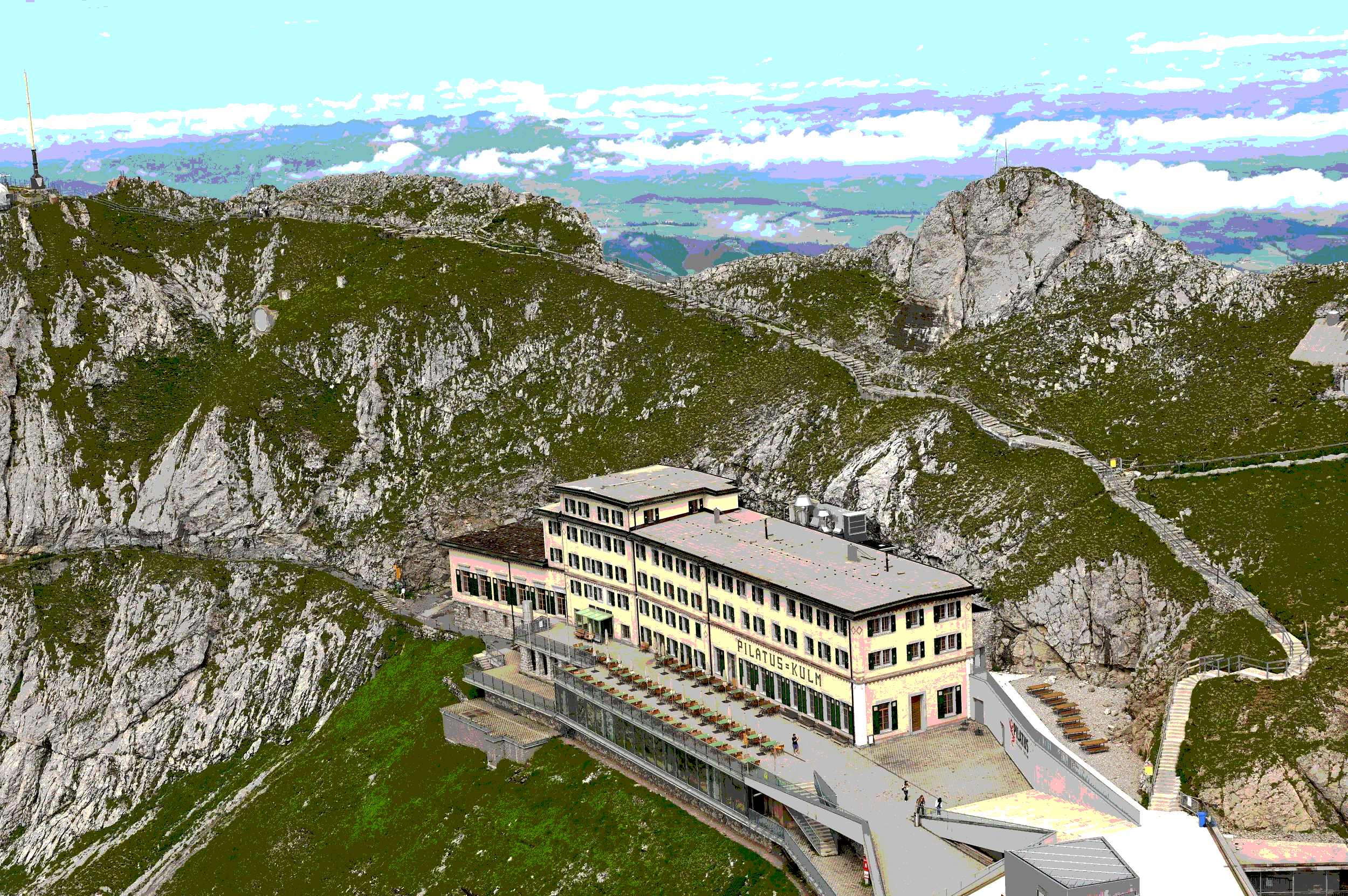 Berg mit Gebäuden, Seilbahn und Wanderwegen, im Hintergrund Berge und Himmel mit Wolken.