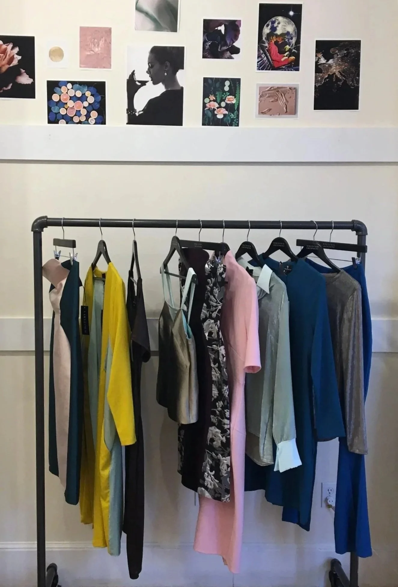 Clothing rack with various dresses and tops in different colors and styles, set in front of a wall decorated with various art prints and images.