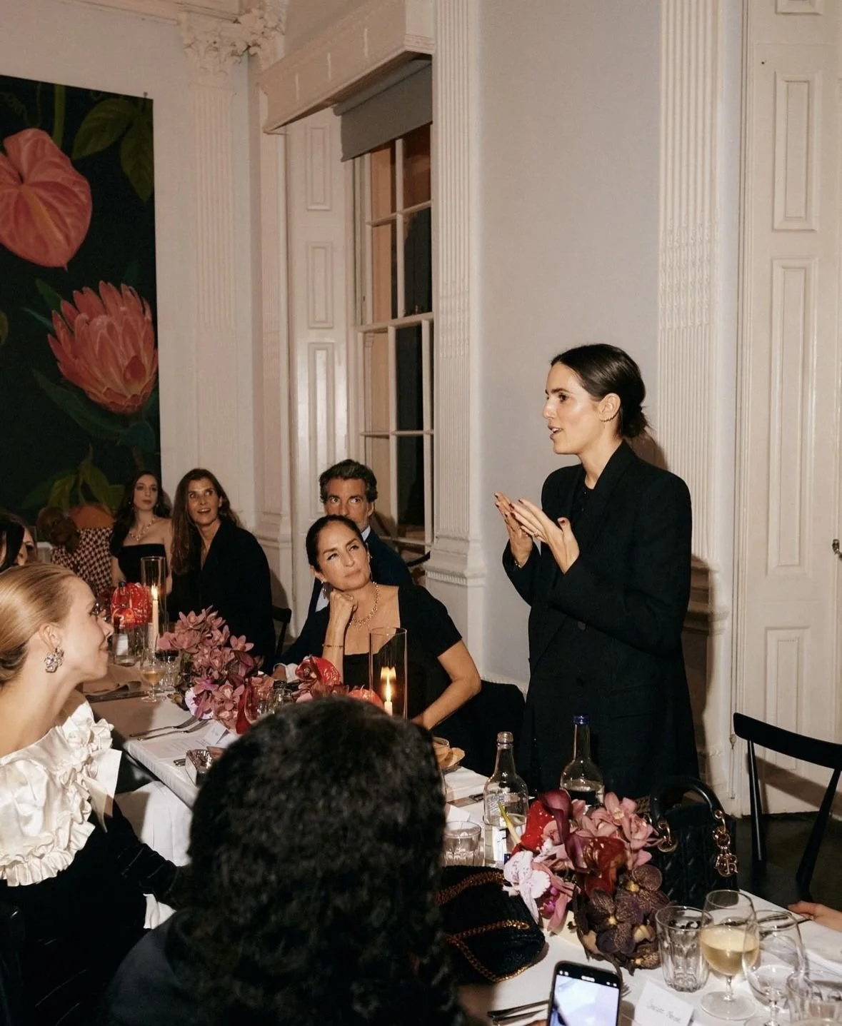 A woman in a black suit giving a speech to seated guests at a formal dinner party in an elegant room with white walls and large windows. The table is decorated with flowers and candles.
