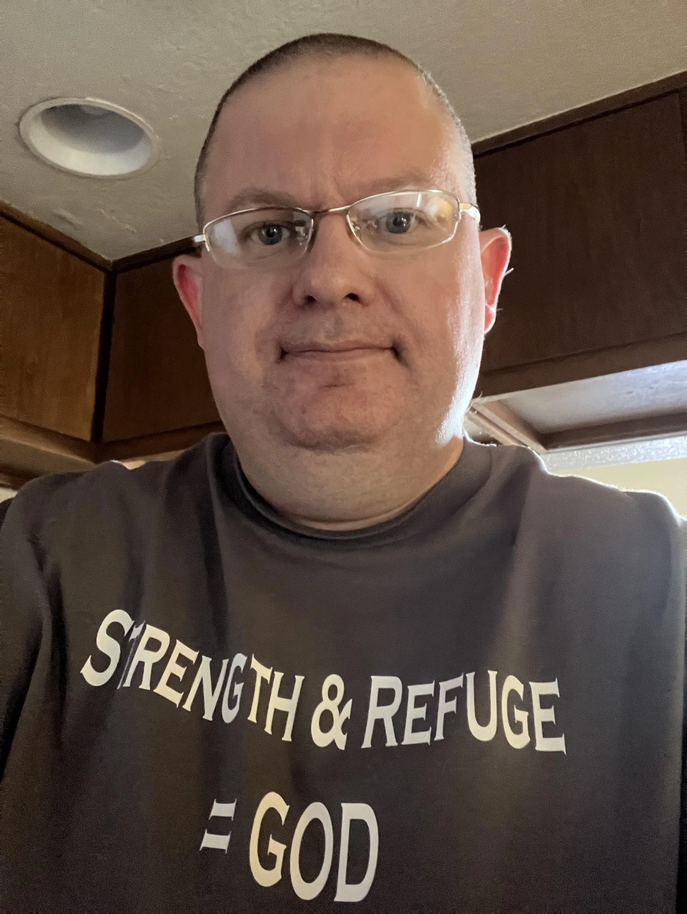 Close-up of a man wearing glasses and a black T-shirt with white text that reads 'Strength & Refuge.'