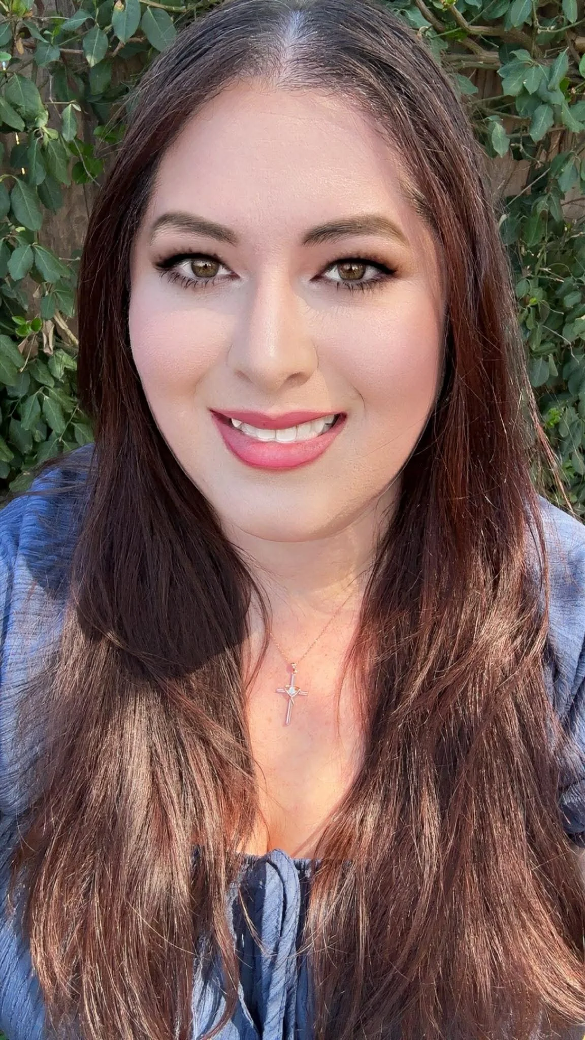 A woman with long brown hair, light skin, and hazel eyes smiling in front of green leafy background, wearing a cross necklace and a blue top.