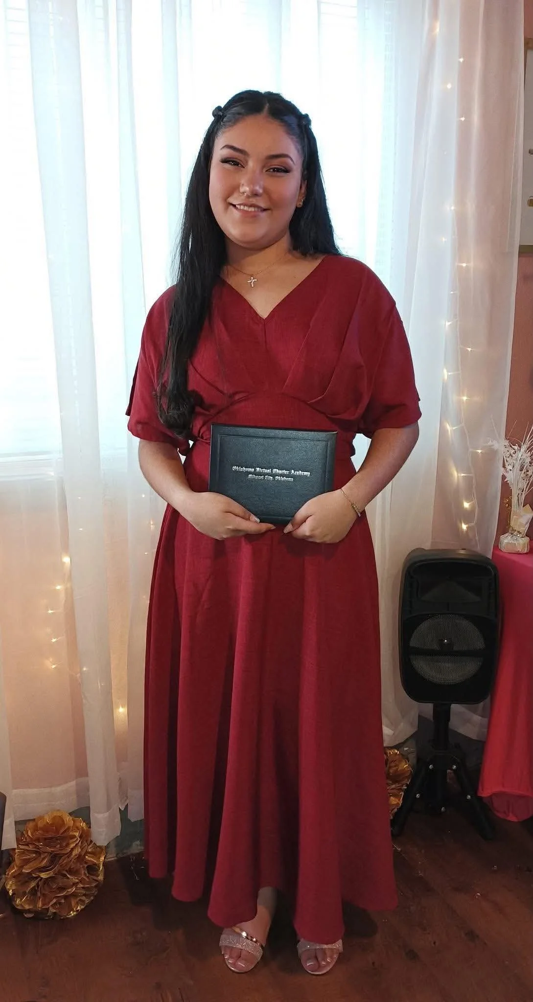 A young woman in a red dress holding a black diploma cover at a graduation ceremony with decorative curtains and fairy lights behind her.