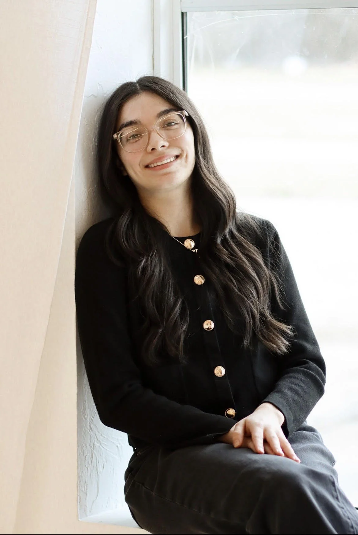 A young woman with long dark hair, glasses, and a black button-up sweater, sitting by a window and smiling at the camera.